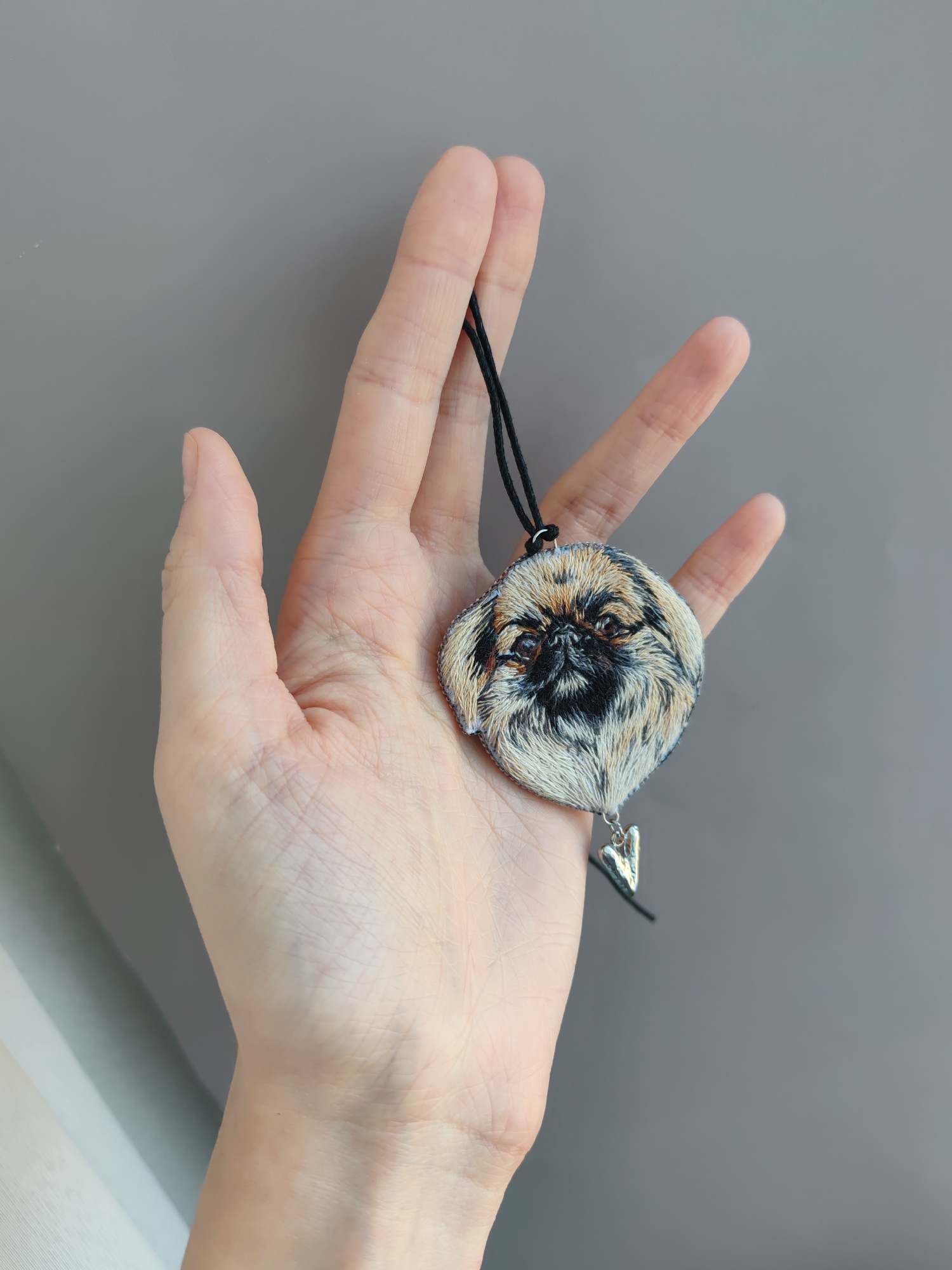 A hand holds a Pekingese dog pendant on a black cord against a grey background. The pendant is detailed with embroidered fur and features a small silver heart charm. The photograph appears to be taken indoors, with a focus on the pendant and the hand holding it. There is no indication that this photo was taken in Ust-Ilimsk, Russia.