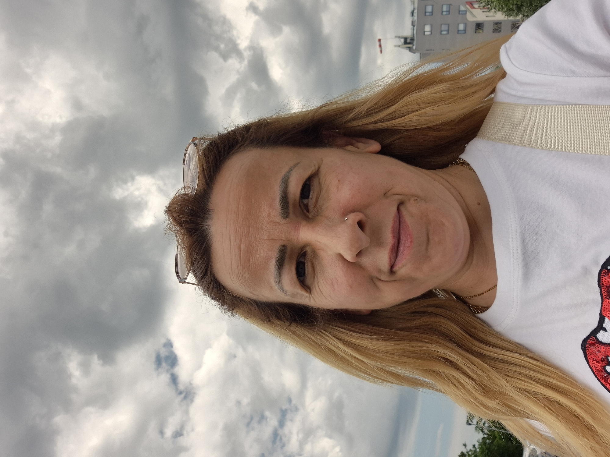 A woman with blonde hair and glasses perched on her head takes a selfie outdoors under a cloudy sky. She is wearing a white t-shirt with a red and black sequin design on the front. In the background, there is a multi-story building.