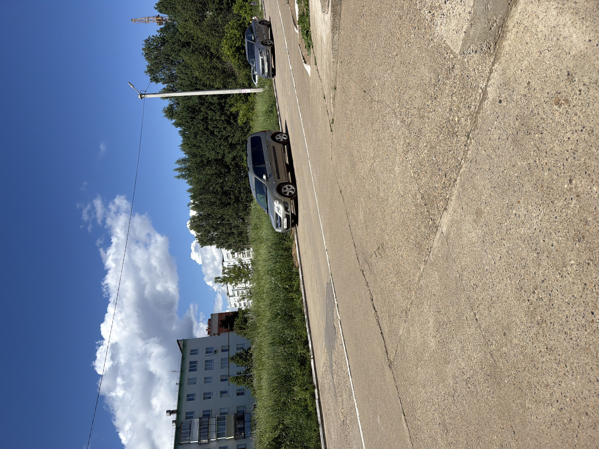 This is a medium-angle, ground-level, daylight, eye-level shot of a street in Chaikovsky, Russia. The scene captures a residential area with a gray asphalt road in the foreground, showing cracks and patches. To the left, a tall, light-colored apartment building with numerous windows stands behind a dense field of green grass. Further to the right, two cars are visible on the road: a silver minivan in the left lane and a silver sedan in the right lane. Beyond the cars, a line of dense green trees forms a natural barrier, and a tall utility pole with a lamp is positioned in front of the trees. In the far background, a construction crane is visible against a bright blue sky with fluffy white clouds. The overall impression is one of a sunny day in a moderately developed urban environment.