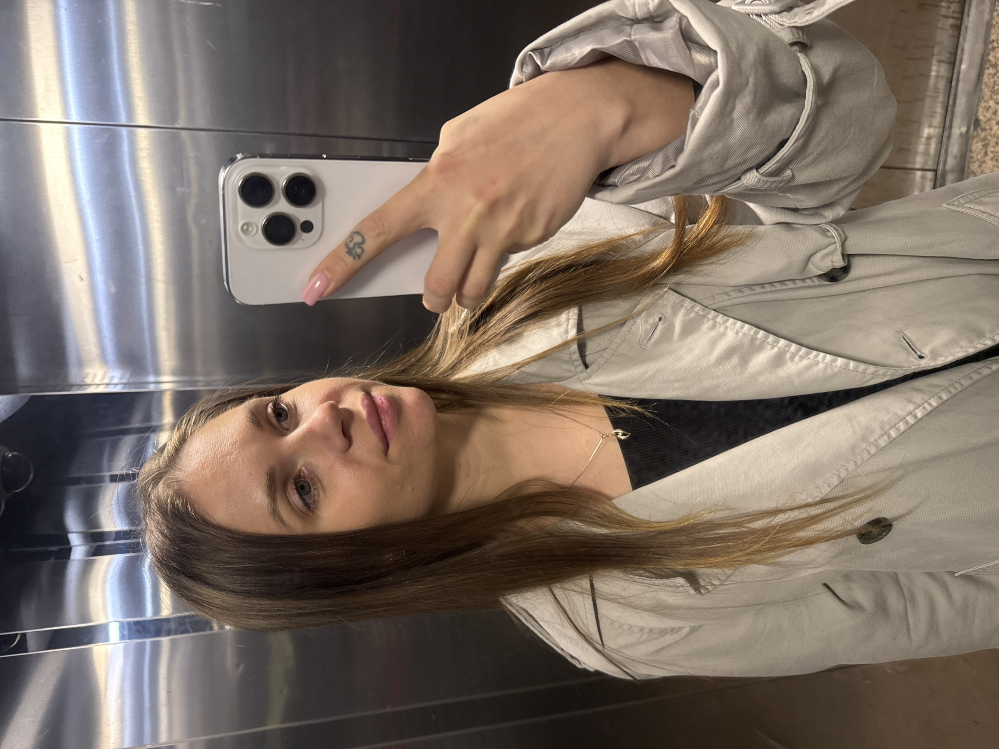 A young woman takes a selfie in a stainless steel elevator. She is fair-skinned with light brown hair, wearing a light gray trench coat over a black top and a gold necklace. Her phone is a white iPhone 13 Pro or newer, and she has a small tattoo on her index finger. The location is assumed to be Batumi, Georgia.