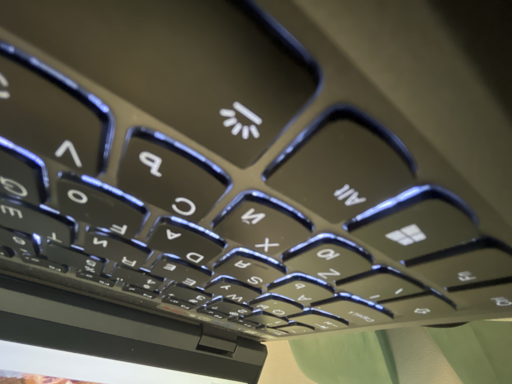 Here is a detailed description of the image:
**Scene:** Close-up view of a laptop keyboard with illuminated keys.
**Objects:**
* Laptop keyboard: Features black keys with white lettering and symbols.
* Illumination: The keys are backlit with a blue/white light that outlines the edges of each key and highlights the characters.
* Key symbols: Visible keys include letters, numbers, and function symbols such as a backlight icon, the Windows logo, "Alt", "Capslk", etc.
**Environment:**
* The background is out of focus, showing a hint of light and possibly a wall.
**Perspective:**
* The image is taken from a low angle, looking up at the keyboard. The keyboard is tilted relative to the viewer, causing perspective distortion.
**Impression:**
* The photo highlights the backlit keyboard, giving it a modern and functional aesthetic.