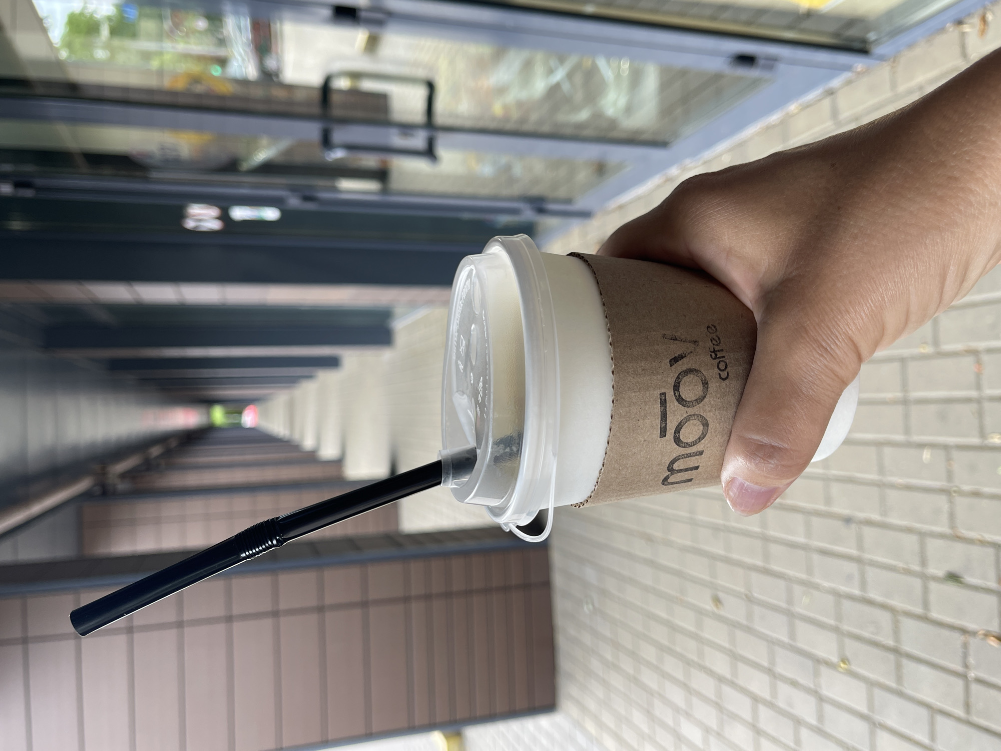 A person's hand holds a paper coffee cup with a brown cardboard sleeve. The sleeve has the logo "moov coffee" printed on it. A black straw extends from the clear plastic lid of the cup. The background shows a long, covered walkway with brown tiled walls and a light-colored tiled floor. To the right, there are glass doors and windows of a building. The image is taken outdoors in what appears to be a city setting. The specific location within Kaliningrad, Russia, cannot be determined from the visual information provided.