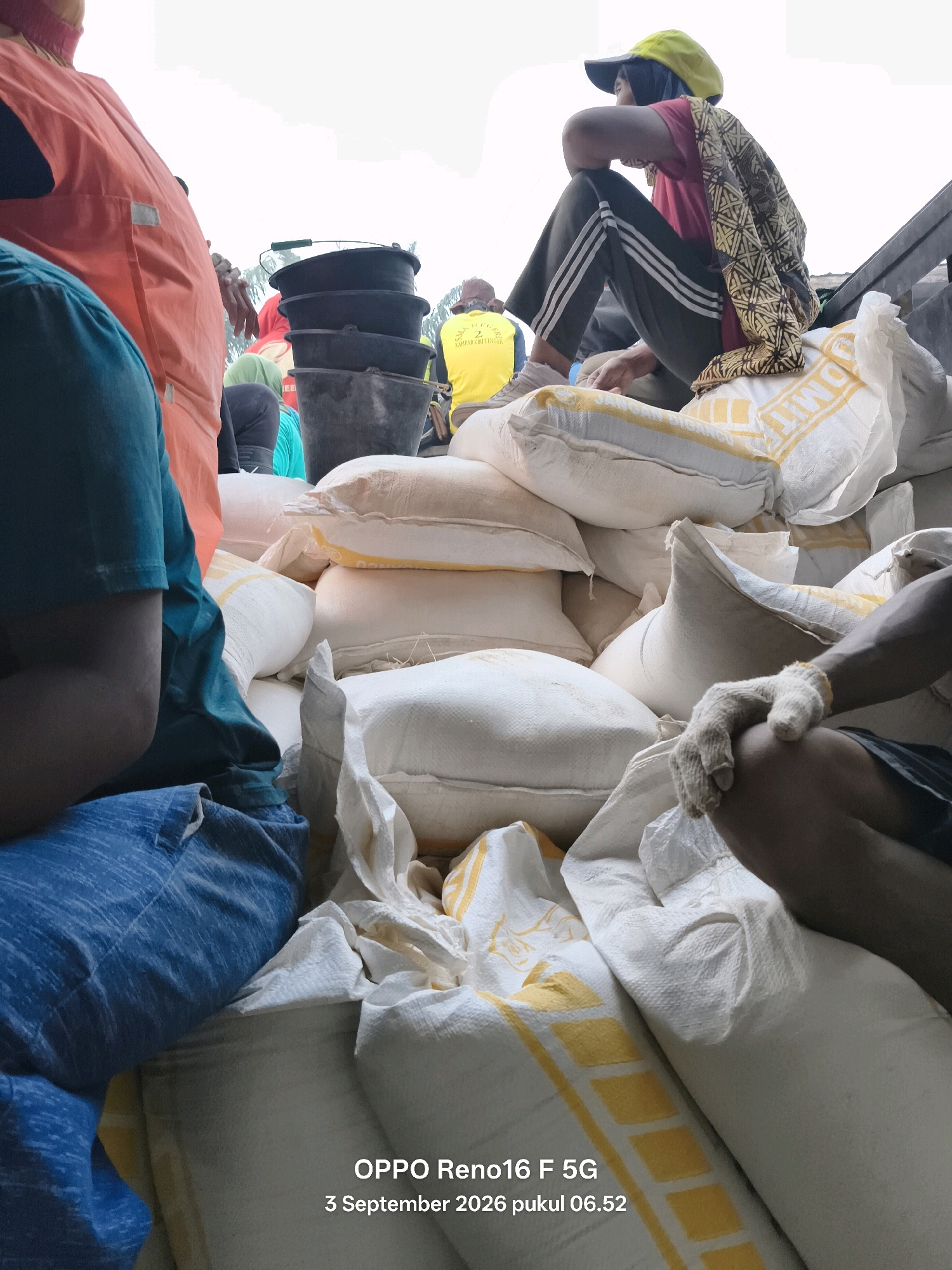 In the city of Penghidupan, Indonesia, on September 3, 2026, at 06:52, several individuals are engaged in activities around a large pile of white sacks, likely containing agricultural produce or similar goods. One person, wearing an orange vest and blue trousers, is partially visible on the left. Another individual, sporting a yellow cap and a red shirt, sits atop some sacks, draped in a patterned fabric. Further back, another person in a yellow shirt is seen. A stack of black buckets is also present in the background. The scene appears to be outdoors, possibly a loading or unloading area, with daylight illuminating the surroundings. The image is a photograph with a timestamp indicating the date and time of capture.