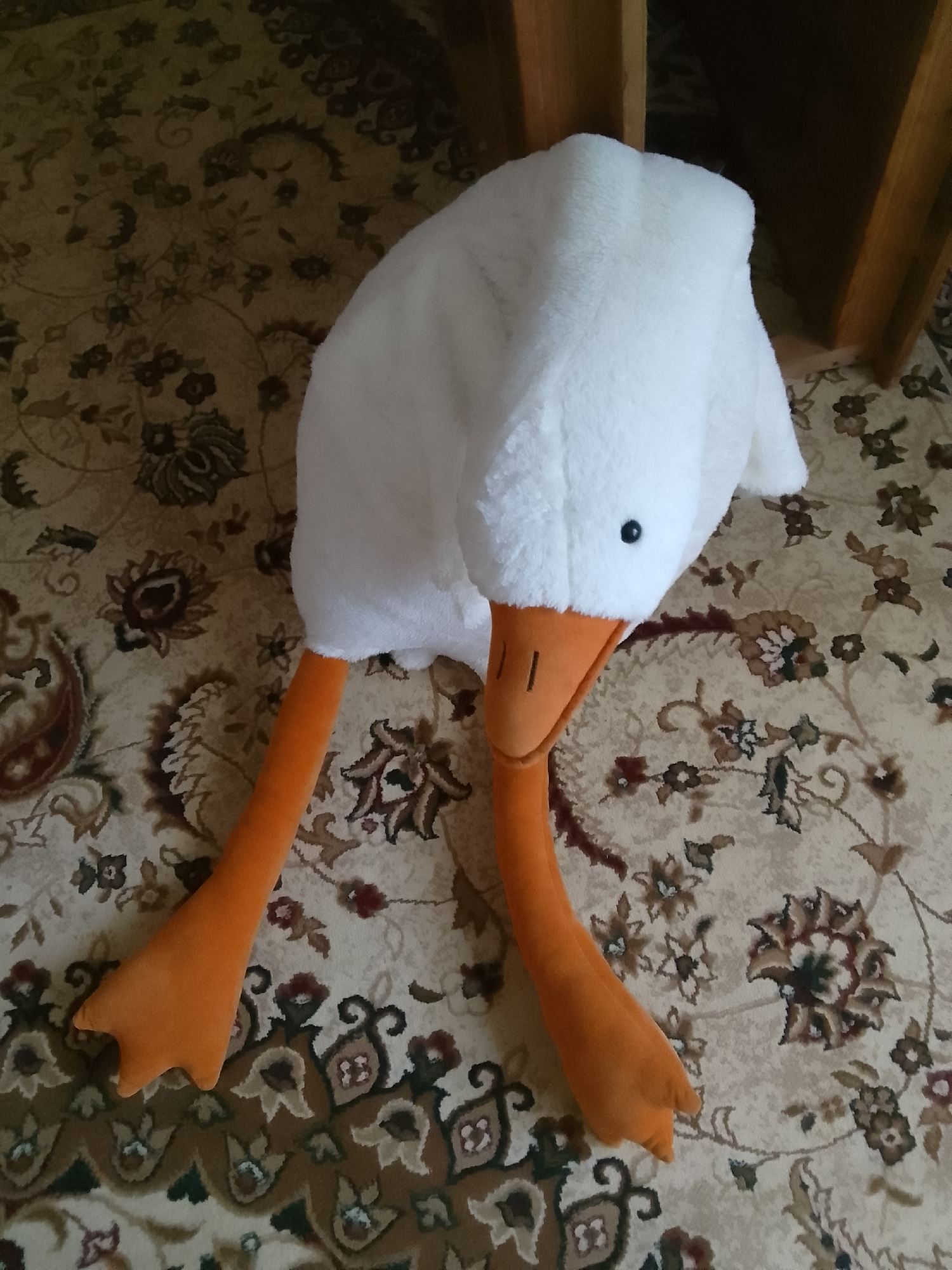 A close-up, overhead shot depicts a novelty plush hat shaped like a goose. The hat is predominantly white with a soft, fluffy texture, featuring a black button eye and an orange beak with two dark lines representing nostrils. Attached to the hat are two long, orange, stuffed legs, each ending in a webbed foot. The goose hat is resting on a patterned rug. The rug has a light beige background with intricate floral and paisley motifs in shades of brown, dark red, and green. The scene appears to be indoors, likely in a room, with a wooden furniture piece visible in the upper right background, suggesting it is part of a home. The lighting is soft and natural, casting faint shadows. The specific location in Taxtakópir, Uzbekistan, is not visually evident from the provided image.