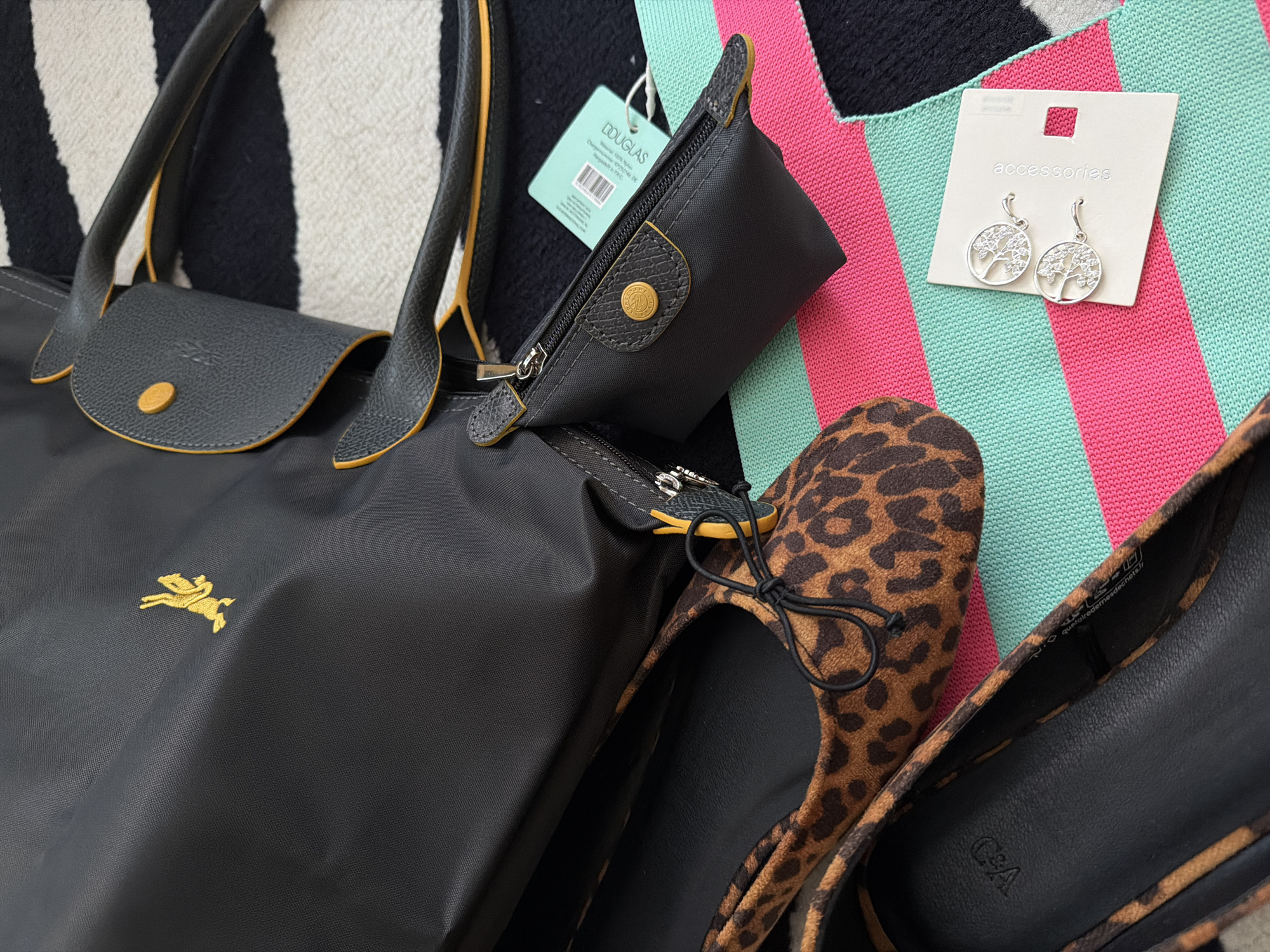 The image displays a collection of personal accessories arranged as a flat lay on a textile surface. The background is a striped fabric featuring wide horizontal bands of black, white, mint green, and hot pink.
Prominently featured is a black nylon tote bag with black leather handles and trim, accented with yellow piping and stitching. A gold-embroidered leaping rabbit logo is visible on the bag's exterior. Adjacent to the tote, a smaller, matching black nylon zippered pouch with a black leather flap and a yellow snap button is present. This pouch also exhibits yellow accents. A light blue-green retail tag, partially obscuring a barcode and text, displays the word "DOUGLAS."
In the lower left quadrant, a pair of leopard print ballet flats with black interiors and black string bows are displayed. The interior sole of one shoe clearly shows the "C&A" brand logo. To the right of the shoes, a pair of silver-colored, circular earrings are presented on a white cardboard backing. The earrings feature an intricate openwork design containing small, clear stones, and the backing is labeled "accessories."
The scene is an indoor setting. No specific geographical landmarks or retail store interiors are visible to precisely identify a location within Perm, Russia; however, the presence of the "C&A" and "DOUGLAS" branded items indicates an association with international retail chains that historically had a presence in the region.
