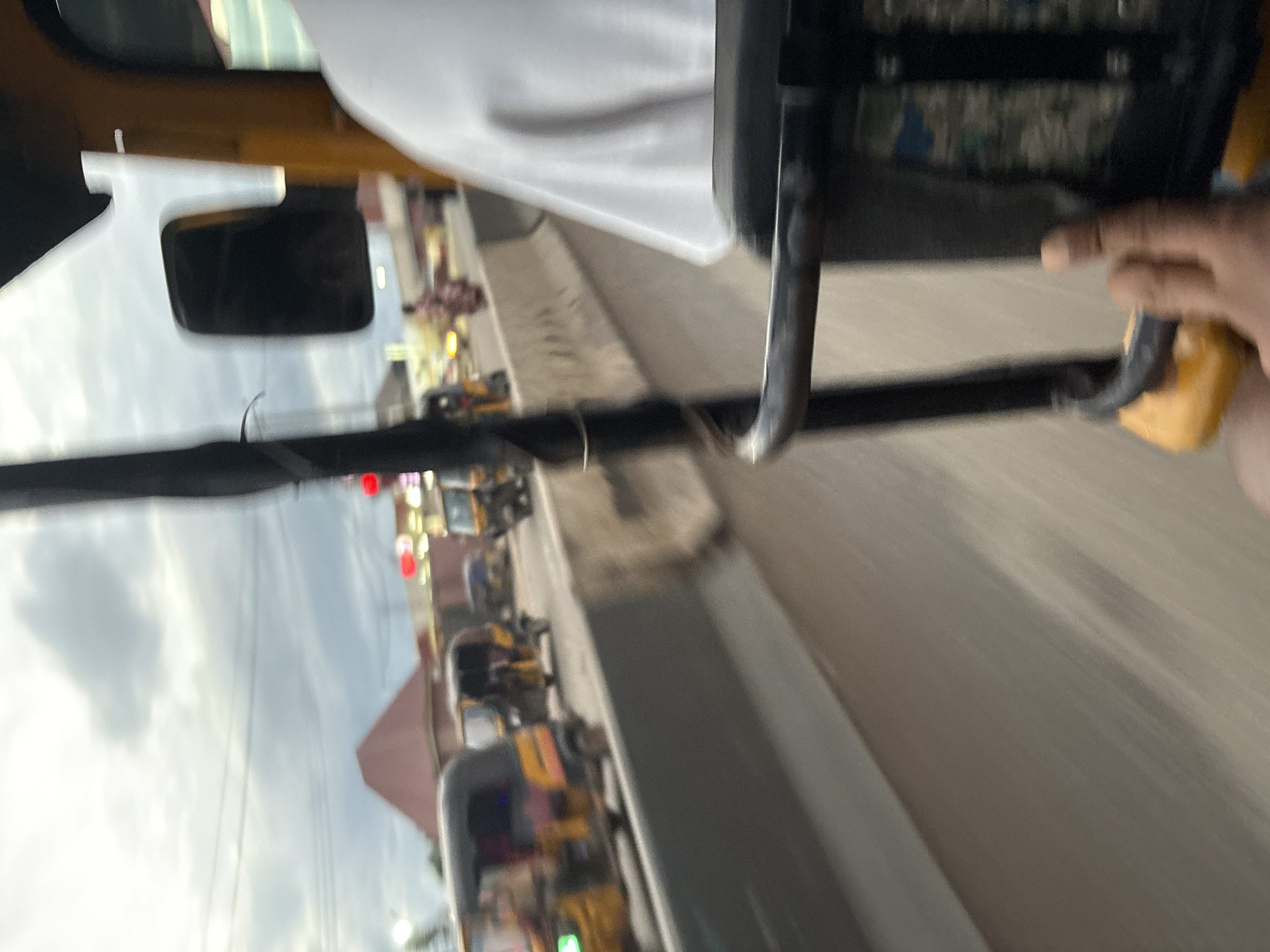 The image captures a motion-blurred view from inside a yellow auto-rickshaw in Onitsha, Nigeria. The foreground shows a portion of the auto-rickshaw's frame and a hand. In the middle ground, a traffic barrier separates the lane of auto-rickshaws from oncoming traffic. Several other auto-rickshaws, also yellow, are visible on the road, some with passengers. Buildings and structures are present in the background, and the sky appears cloudy. The scene suggests a busy street in Onitsha, likely during daytime.