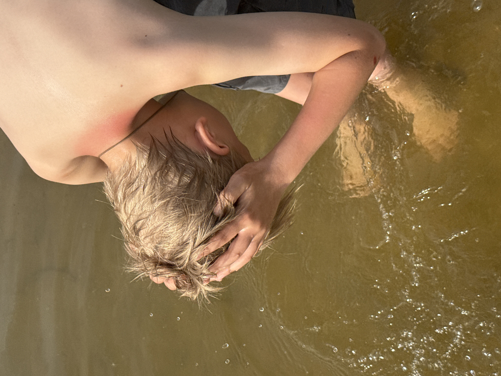 A boy is submerged in murky water, possibly in Novosinkovo, Russia. He has light blonde hair and is partially submerged. He rests his head in his hands, which are also in the water. He appears to have a sunburn on his back. His feet are also submerged in the water.