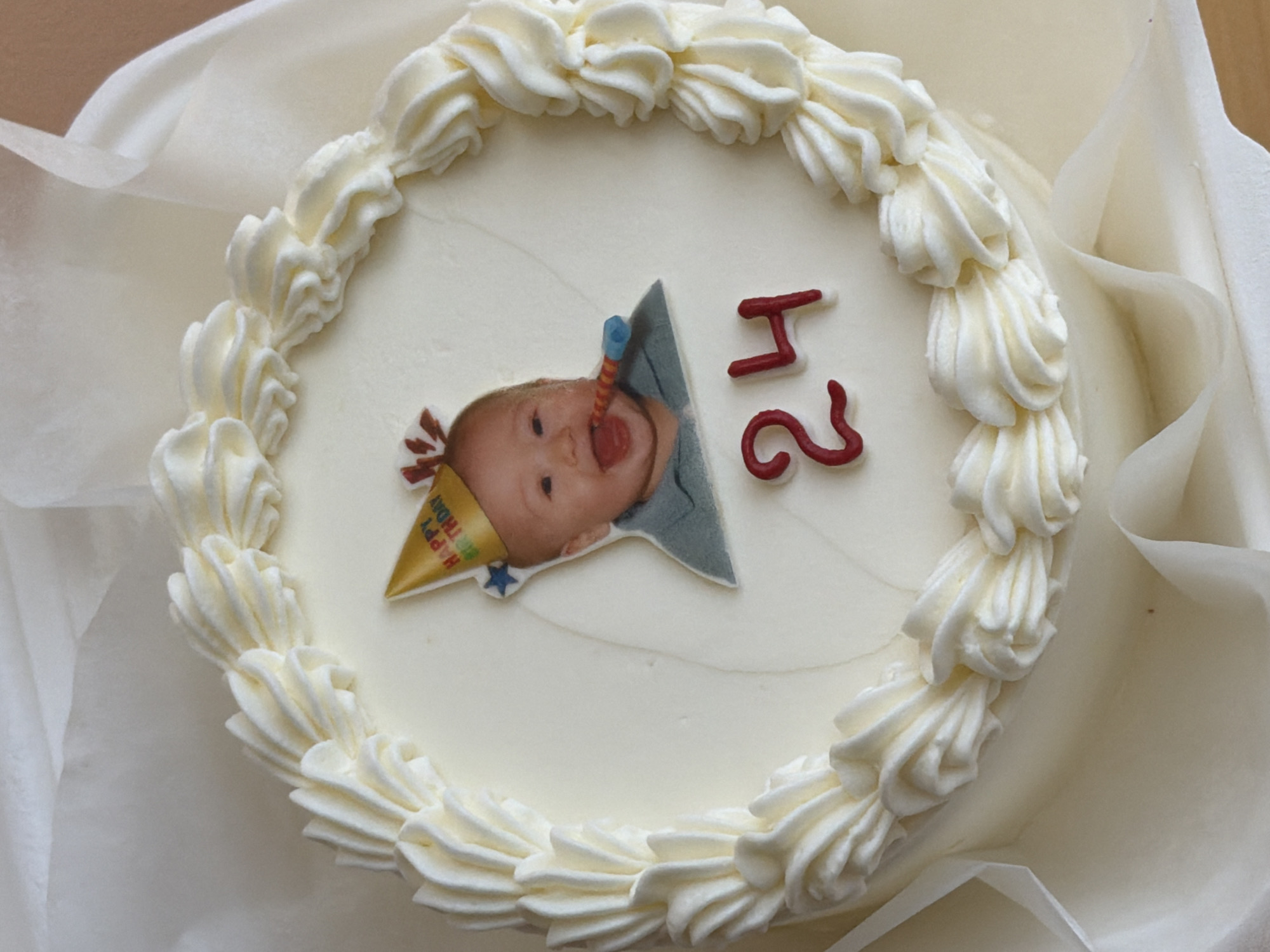 A top-down shot depicts a white birthday cake, likely homemade, decorated with white frosting in a circular pattern around the edge. A printed image of a baby wearing a birthday hat and blowing a party horn is centrally placed. The number "24" is formed with red frosting, positioned beside the baby image. The cake sits on parchment paper inside a white box. The location is not discernible from the image; thus, the Tel-Aviv reference cannot be confirmed.