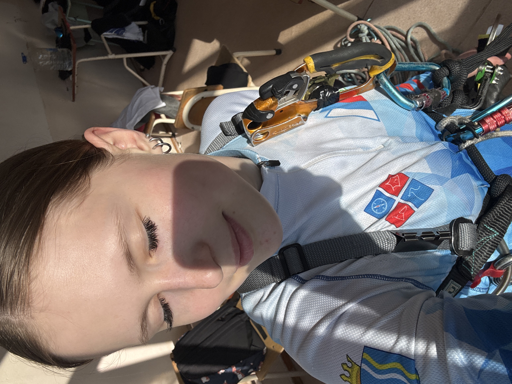 The image shows a young woman lying down and wearing a climbing harness with climbing equipment attached. The harness has a blue and white patterned material on the front, and the pattern includes a logo with the image of waves with a crown above them, and another logo that includes red and blue squares and a compass. The equipment is various climbing gear, including carabiners, ropes, and a mechanical rope grab. The woman is fair-skinned and has dark hair and long eyelashes. The background includes a chair and some bags. While logos are shown, it is not possible to discern the location in Berezniki, Russia.