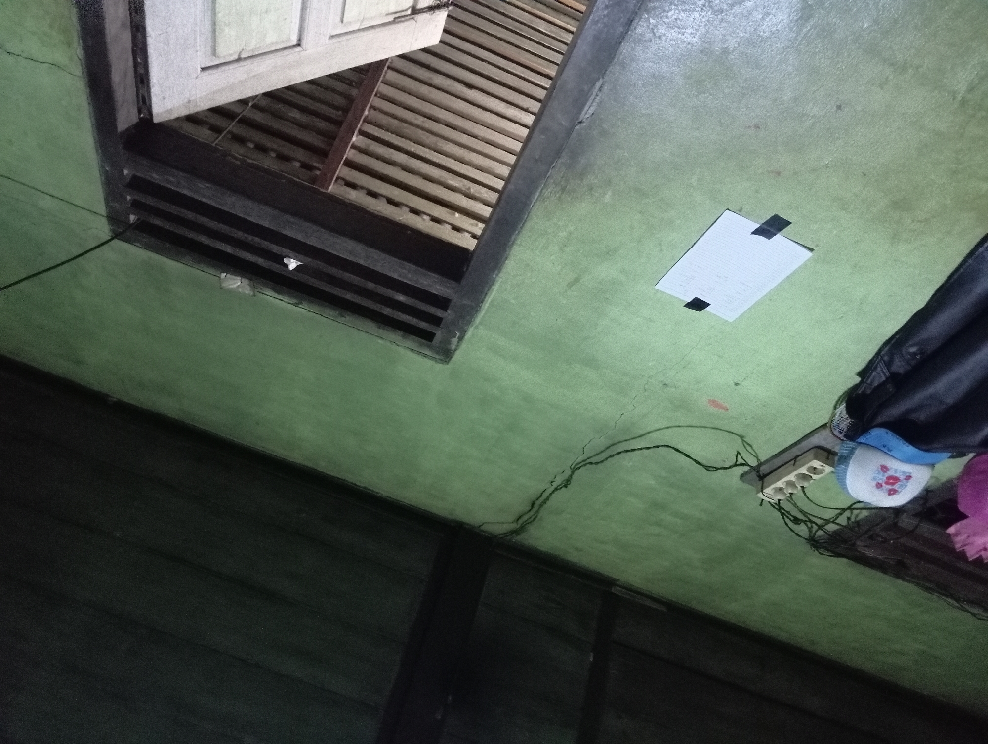 The image depicts an interior scene in Rejosari, Indonesia, with a light green wall dominating the frame. In the lower left, a black jacket is draped over a white and blue trucker hat. To the right of the jacket and hat, a multi-outlet power strip with three sockets is plugged into a wall socket. Cables snake across the wall from the power strip. In the center of the wall, a piece of white paper is taped down with black tape. To the right, a doorway with wooden louvers is visible, leading to a darker interior. The ceiling, made of dark wooden planks, is visible in the upper portion of the image.