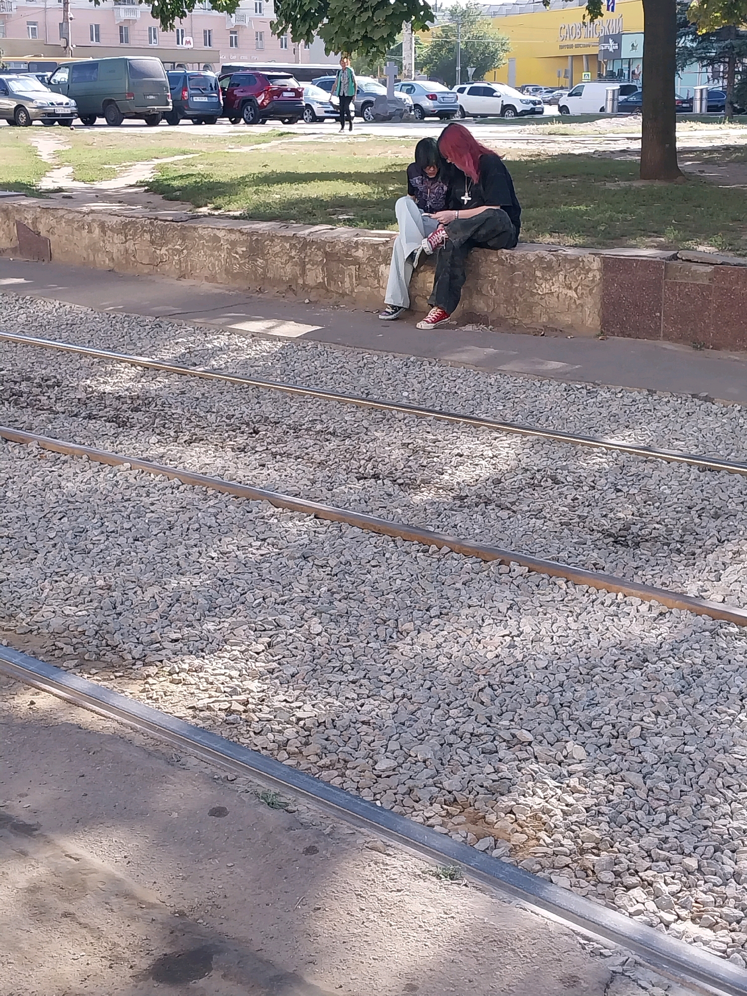 Two individuals are seated on a concrete ledge next to a set of tram tracks. The person on the left has dark hair and is wearing a gray top and light-colored pants with red and white shoes. The person on the right has long, reddish hair and is wearing a black t-shirt and dark pants with red and white shoes. They are both looking down at something in their hands, appearing to be engaged in a shared activity, possibly looking at a phone or book. The environment suggests an urban setting with a paved area and a grassy strip beyond the concrete ledge. In the background, buildings and parked vehicles are visible, indicating a street or public area. The tram tracks are comprised of gravel and metal rails.
