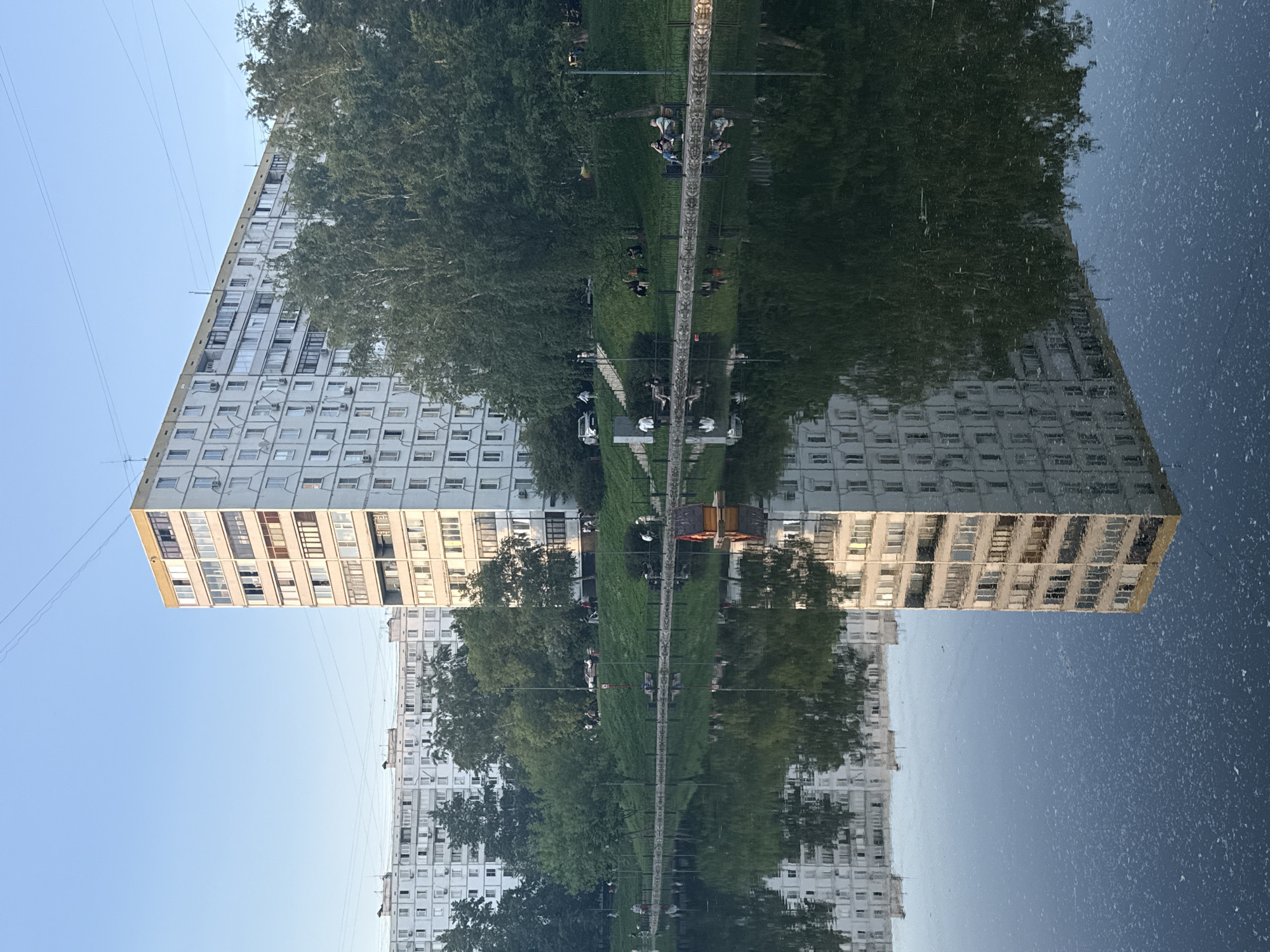 The image depicts a reflection of a building and park area, likely caused by a body of water. The building is a large, white apartment block with numerous windows. A park area is situated next to the building, featuring trees and a walkway. People appear to be sitting on benches or walking along the path. The reflection in the water distorts the image, creating a symmetrical effect. Based on the architecture and general appearance, the location is likely in Moscow, Russia.