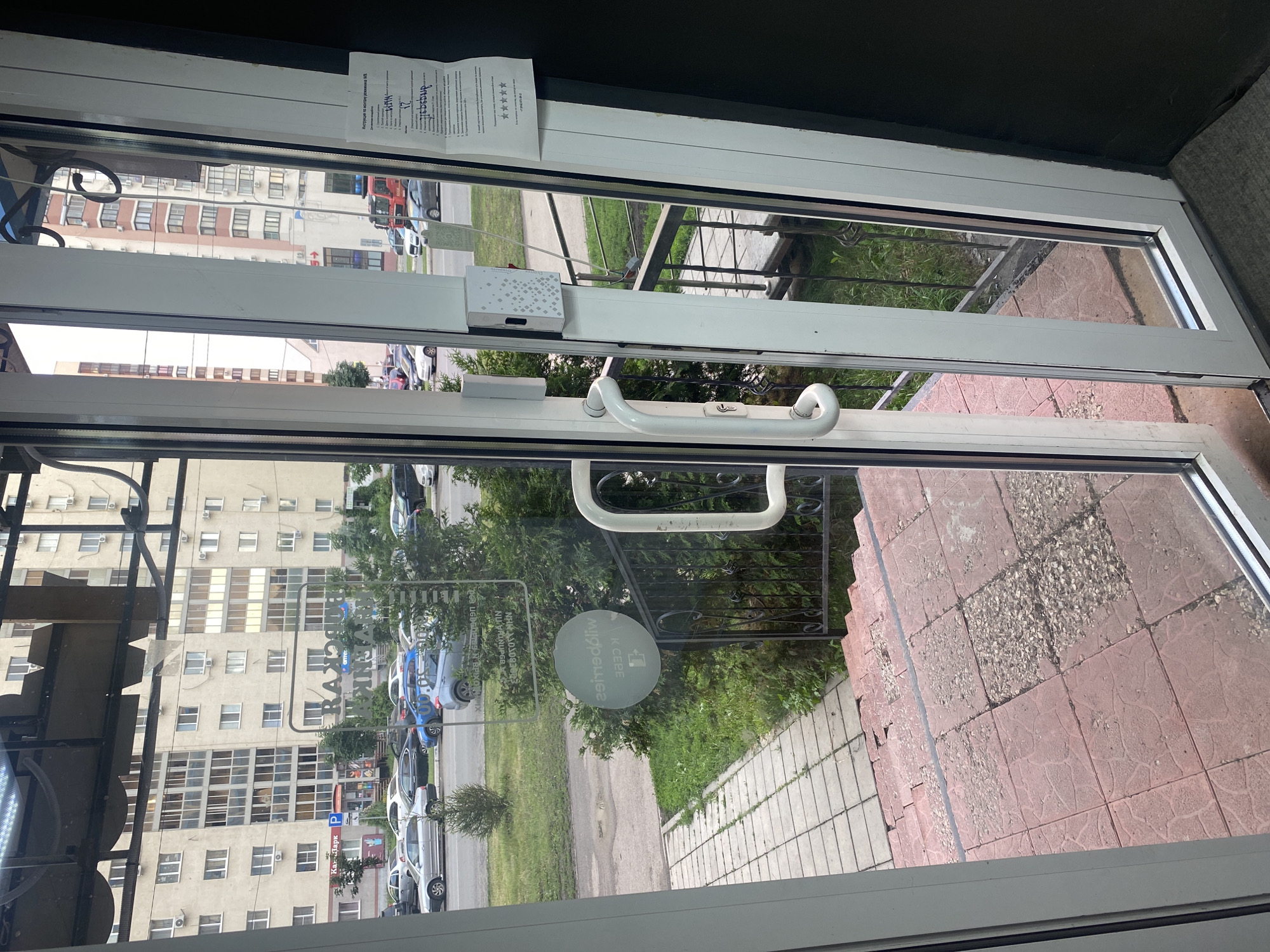 The image shows a street scene in Rozhdestveno, Russia, viewed through a glass door. In the foreground, a white metal door handle is prominent, and to the left, part of a tiled staircase is visible. Outside, a sidewalk leads to a street lined with parked cars. In the background, several apartment buildings are visible, with architectural details typical of urban Russian housing. A red fire truck is parked near one of the buildings. The overall impression is that of a residential urban environment.