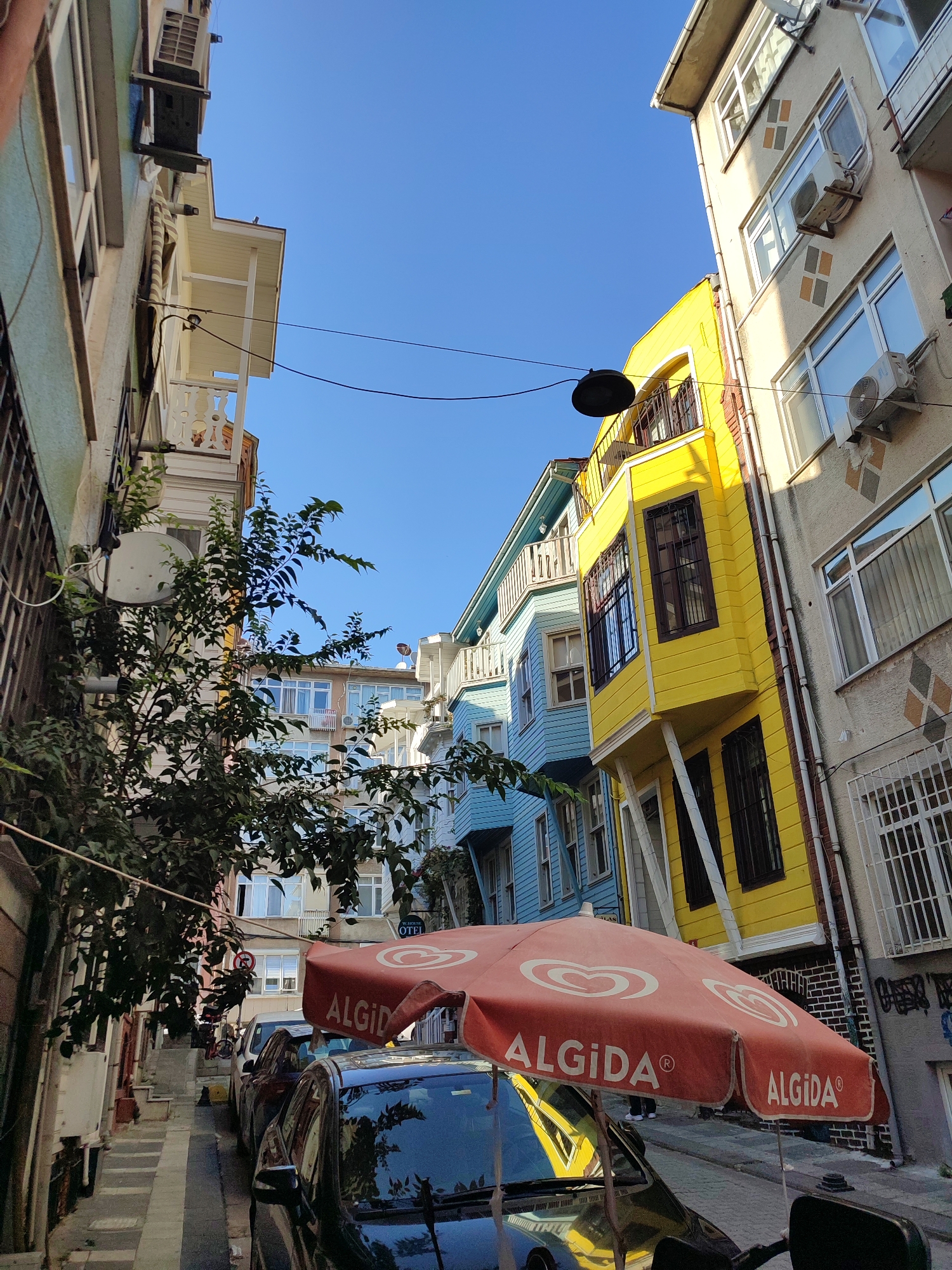 The image depicts a narrow street in Kadıköy, Turkey, lined with colorful buildings under a clear blue sky. A bright yellow building with dark-framed windows and a black metal balcony stands out, adjacent to a light blue building with a similar balcony and white railings. To the right, a pale beige building features multiple windows and air conditioning units. A large, orange umbrella with the "Algida" logo is open over a parked black car, partially obscuring the street. Green foliage from a tree on the left side of the frame extends into the scene, with its leaves and branches creating shadows. In the background, a staircase leads further up the street, with more buildings visible. There are no people actively engaged in interactions, but their presence is implied by the parked cars and the open umbrella. The environment appears to be residential or mixed-use, with elements of urban life such as power lines and satellite dishes present.