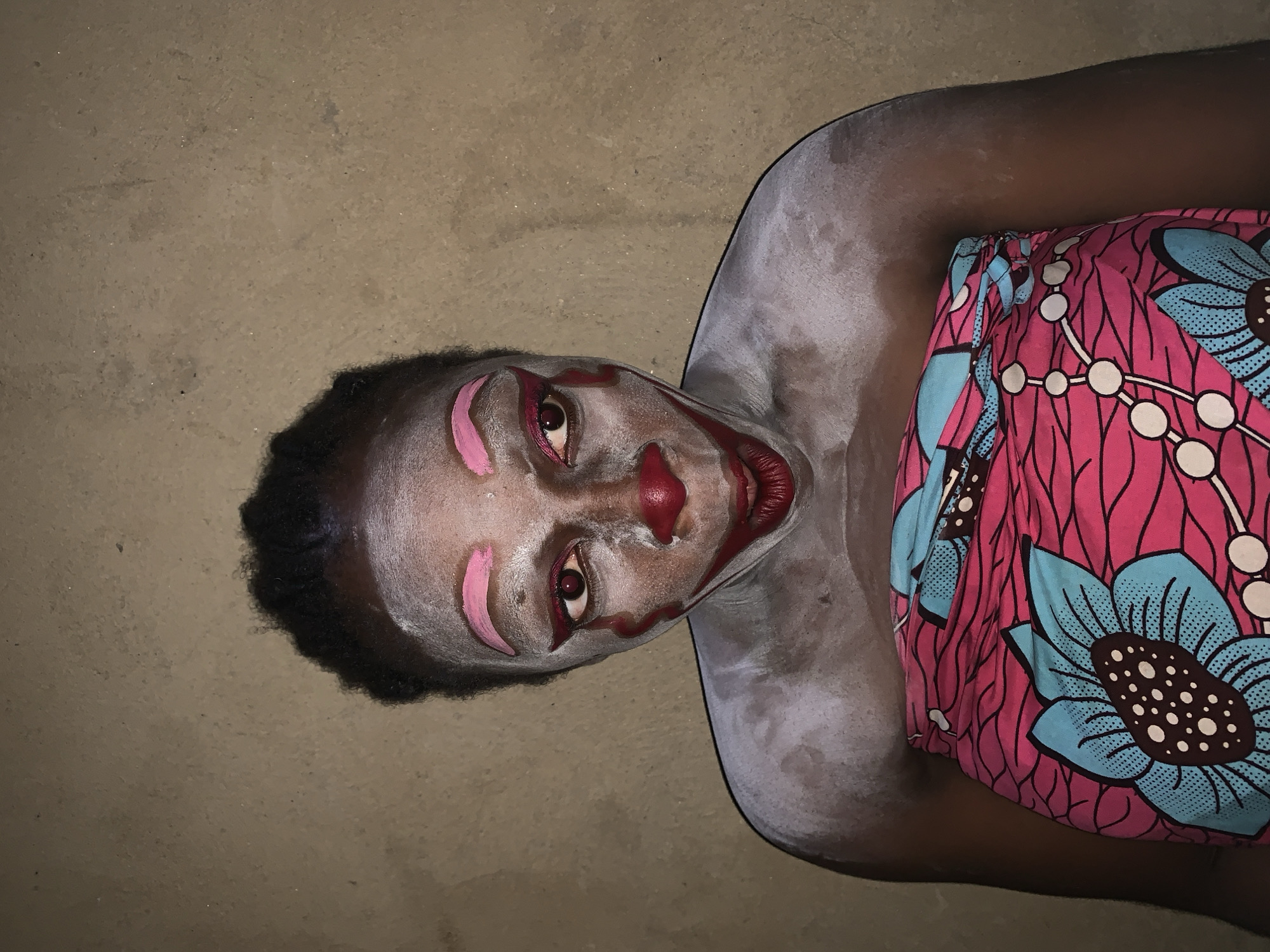 The image shows a person wearing clown-like makeup, with white powder applied to their face and shoulders, and pink and red accents on their eyes, eyebrows, nose, and lips. The person is draped in a brightly colored fabric with a floral pattern. The background is a rough, textured wall, suggesting an indoor or sheltered outdoor environment. The setting is described as the city of Agban, Nigeria.