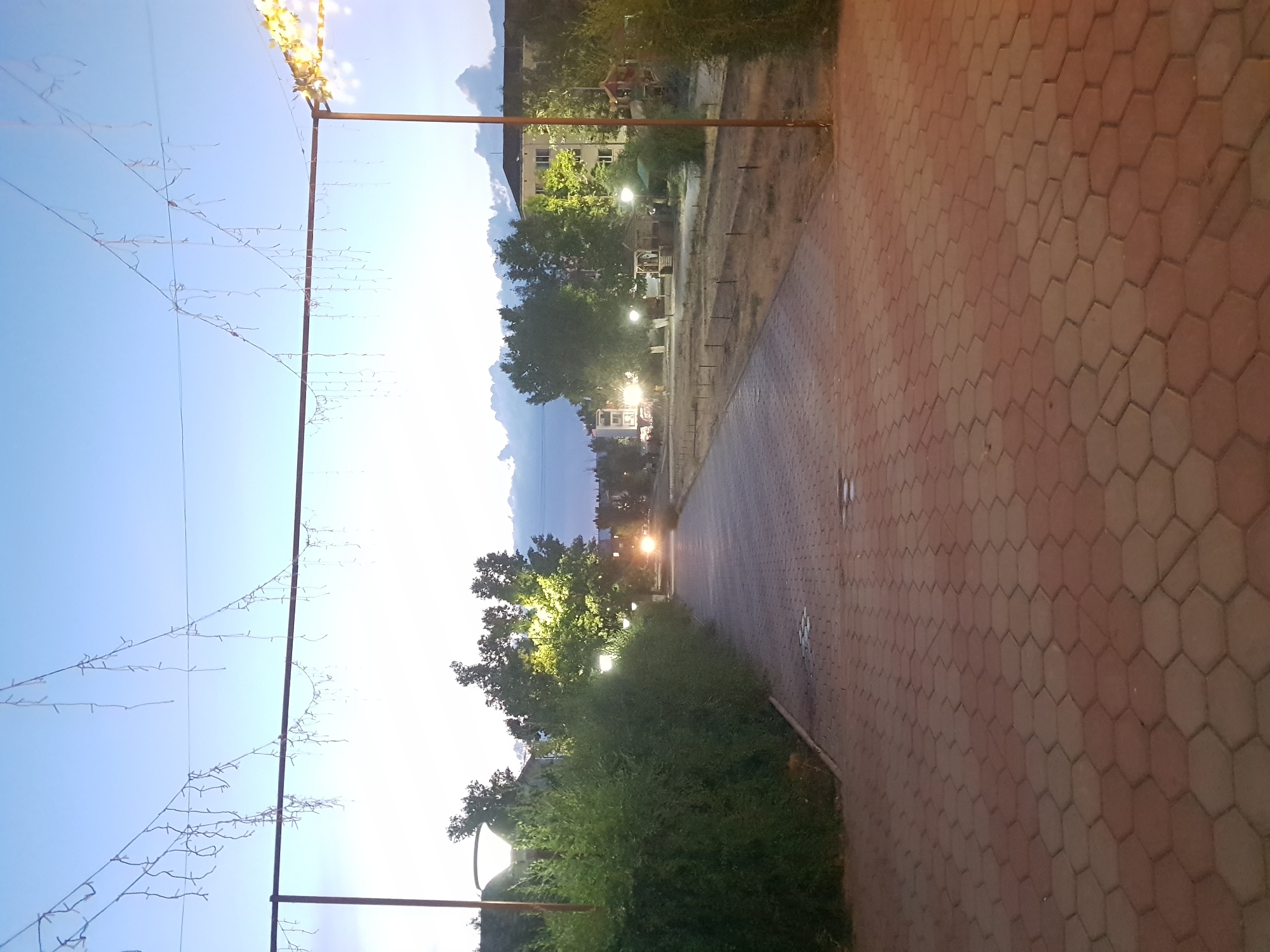 The image depicts a walkway paved with hexagonal reddish-brown bricks, leading into the distance. The walkway is lined on the left by a dense, well-maintained green hedge and on the right by dry, patchy grass and some small saplings. Overhead, two sets of strings with decorative lights are suspended, one set lower and closer, the other higher and further away. The sky is a gradient of pale blue and white, suggesting twilight or a very early morning.
In the distance, a vehicle with illuminated headlights is visible on the walkway. Several trees, some with their leaves lit by nearby streetlights, are scattered throughout the scene. Behind the trees, buildings are visible, including a multi-story apartment building and what appears to be a playground structure. The general atmosphere is calm and serene, with the lights of the walkway and the vehicle providing points of illumination against the fading natural light.
The location is identified as Kurchatov, Kazakhstan. The scene appears to be a public park or a communal area within a residential complex, possibly during a festive season or simply as part of the town's ambiance. There are no people clearly visible in the image.