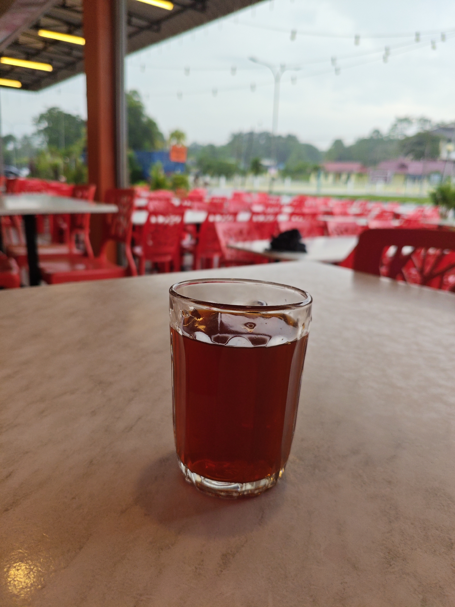 A close-up, eye-level shot focuses on a glass of dark amber liquid, possibly iced tea or coffee, situated on a light brown, speckled table. The glass, made of clear, textured glass, is half-filled with the beverage. The background is blurred but reveals an outdoor seating area with numerous bright red plastic chairs arranged around tables. The scene appears to be a food establishment, possibly a hawker stall or a restaurant, in Kota Tinggi, Malaysia, given the typical outdoor seating arrangements and bright red chairs commonly found in such locations. The lighting suggests it is daytime, with an overcast sky visible in the distance. The overall ambiance is casual and relaxed, hinting at a typical Malaysian roadside eatery.