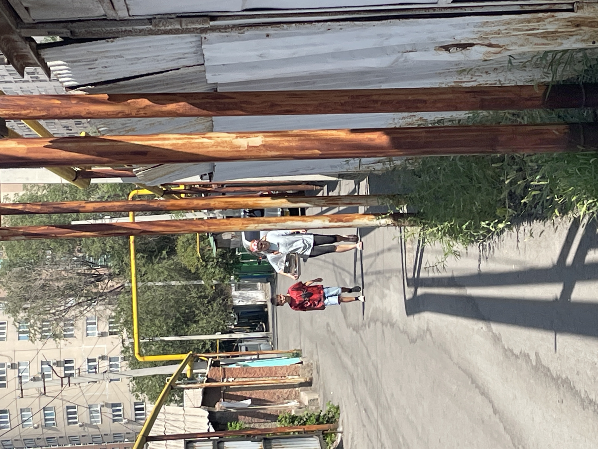 A boy and a woman are walking down a narrow, paved street in Almaty, Kazakhstan. The boy, wearing a red t-shirt and blue shorts, walks slightly ahead of the woman, who is carrying a tray with some items. They are walking away from the camera, down an alleyway lined with buildings and fences. In the background, a tall apartment building and trees are visible. The street is shaded by structures and utility poles, creating long shadows on the ground. The overall scene depicts a typical urban environment with a mix of residential and industrial elements.