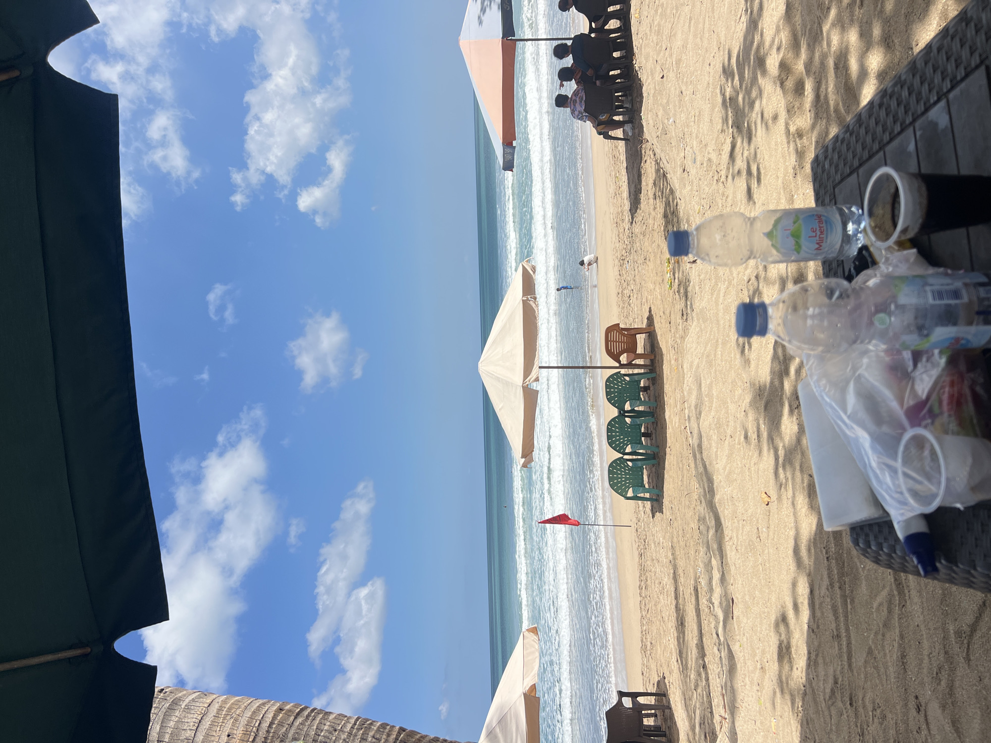 The image depicts a beach scene in Kuta, Indonesia, on a bright, sunny day. In the foreground, a table is set up with two plastic water bottles, one labeled "Le Minerale," and a cup of dark liquid, possibly coffee or soda. A plastic bag containing various items, possibly snacks, is also present. In the middle ground, a sandy beach stretches towards the ocean, with several green plastic chairs lined up and a single brown plastic chair placed slightly apart. A red flag, likely a warning signal, stands on the beach. Two beach umbrellas provide shade, one light-colored and another with orange and gray panels. In the background, people are visible on the beach and in the shallow water. Some individuals are seated under the orange and gray umbrella, and others are playing or wading in the ocean. The sea is calm with gentle waves, and the sky is blue with scattered white clouds. The overall atmosphere suggests a relaxing day at the beach.