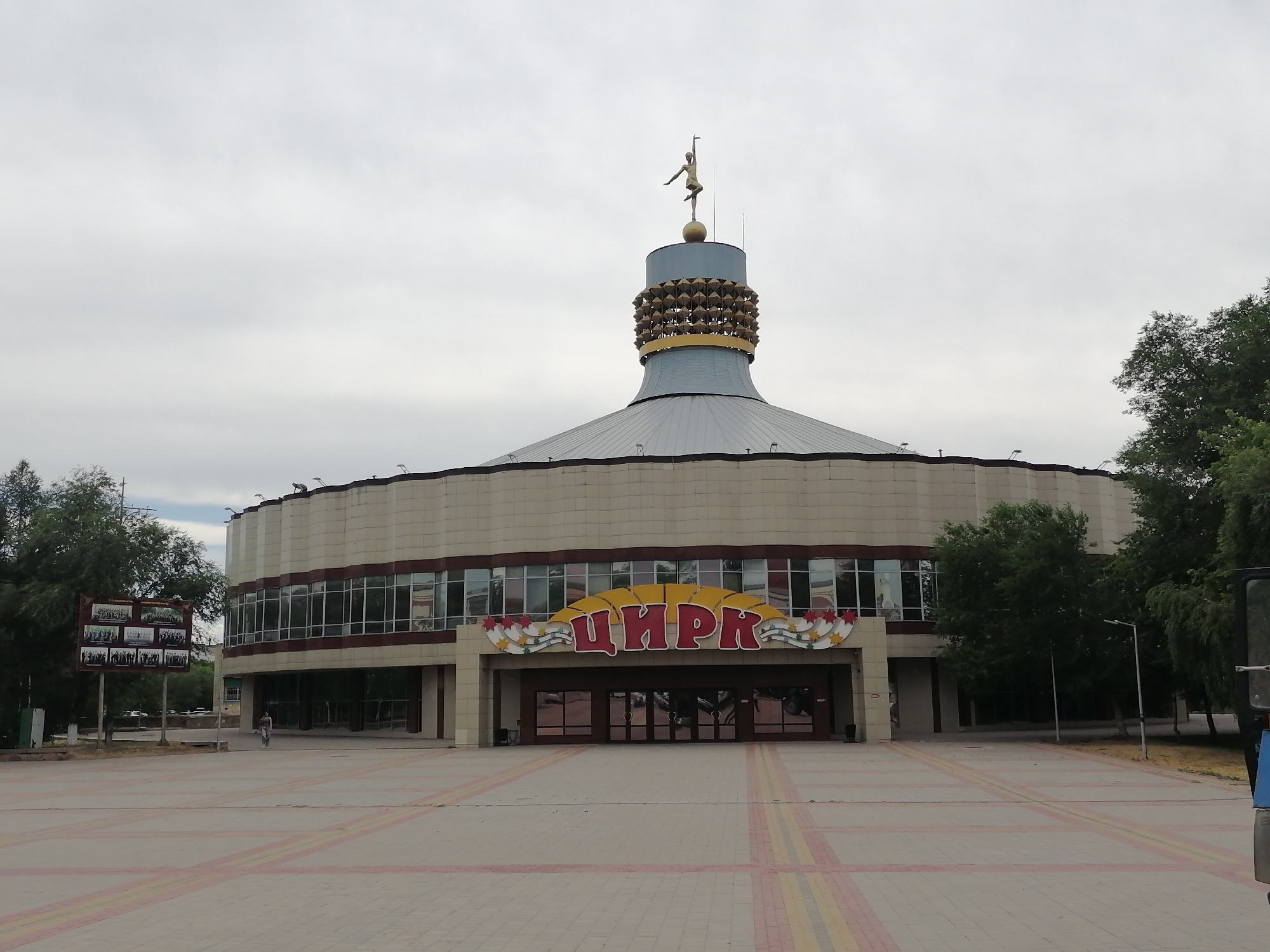 The image shows the Karaganda Circus in Karaganda, Kazakhstan. The circus building is circular with a domed roof and a spire topped by a statue of a woman on a ball. The building is clad in beige panels and has large windows. Above the entrance, there is a large sign with the word "ЦИРК" in red letters within a yellow sunburst design. In front of the building is a paved plaza with a pattern of red and yellow tiles. Trees are visible on either side of the building and in the background. A small number of people are visible in the plaza, and a portion of a blue vehicle is present on the right side of the image. A large display board with photographs is situated to the left of the building. The sky is overcast.