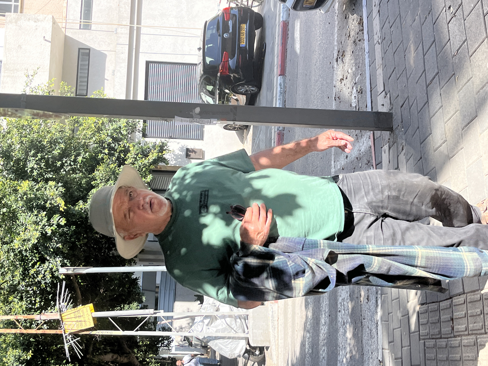 A man wearing a grey hat, green t-shirt, and grey pants stands on a sidewalk in Tel Aviv, Israel. He holds sunglasses in his left hand and a plaid jacket hangs on his right side. Behind him, there is a black car with a license plate number 332-46-201 parked next to a building with closed shutters. A tree is visible to the left of the man. A street lamp is in front of the tree. The sidewalk is paved with grey bricks.