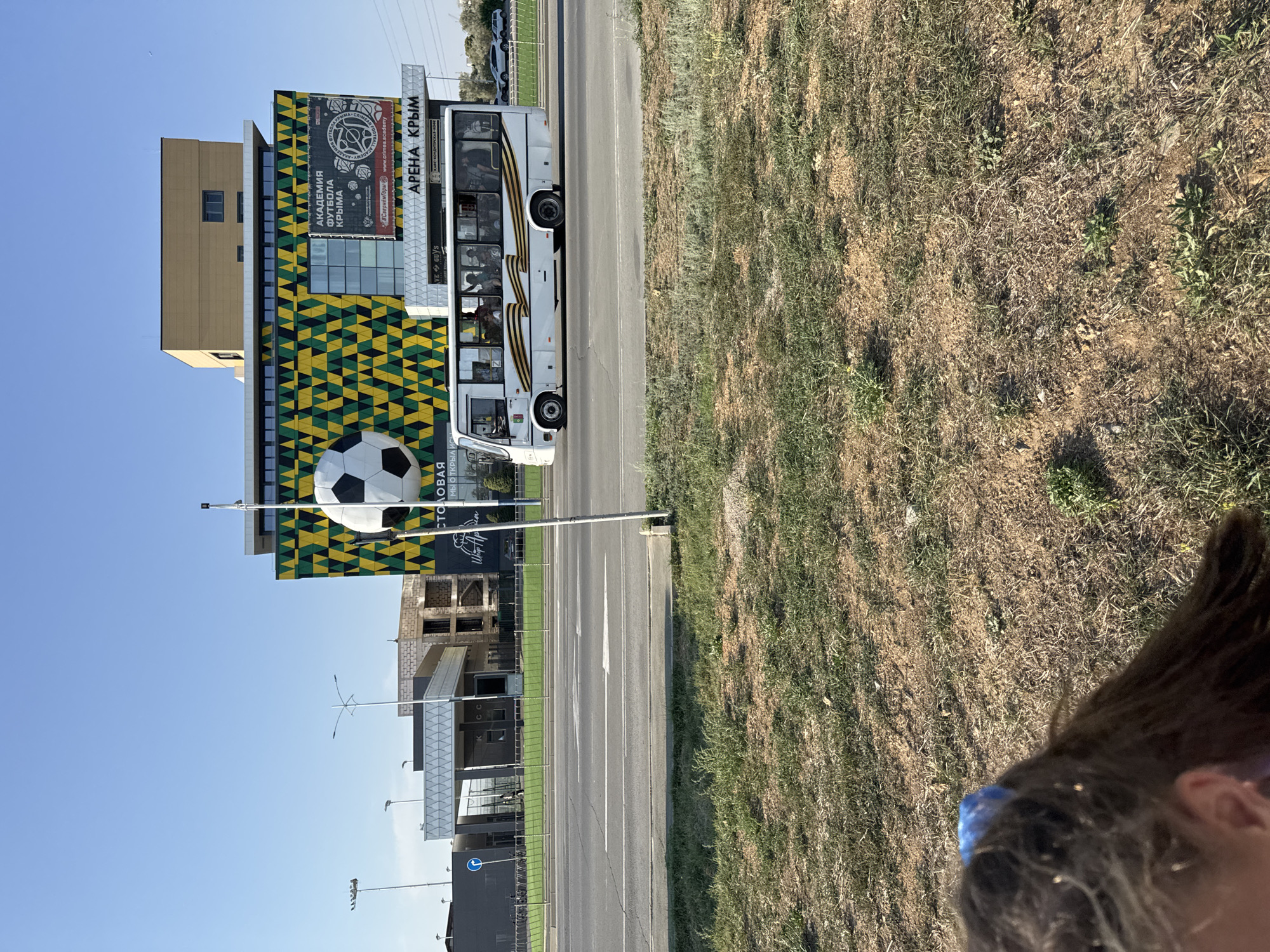 A white bus with a St. George's ribbon decal on its side is driving down a road in front of a building with a large soccer ball sculpture on its facade in Yevpatoriya, Ukraine. The building is a sports complex named "Arena Krim" and features a yellow and green patterned exterior. The bus is filled with passengers, and the scene is set under a clear blue sky with sparse vegetation in the foreground.