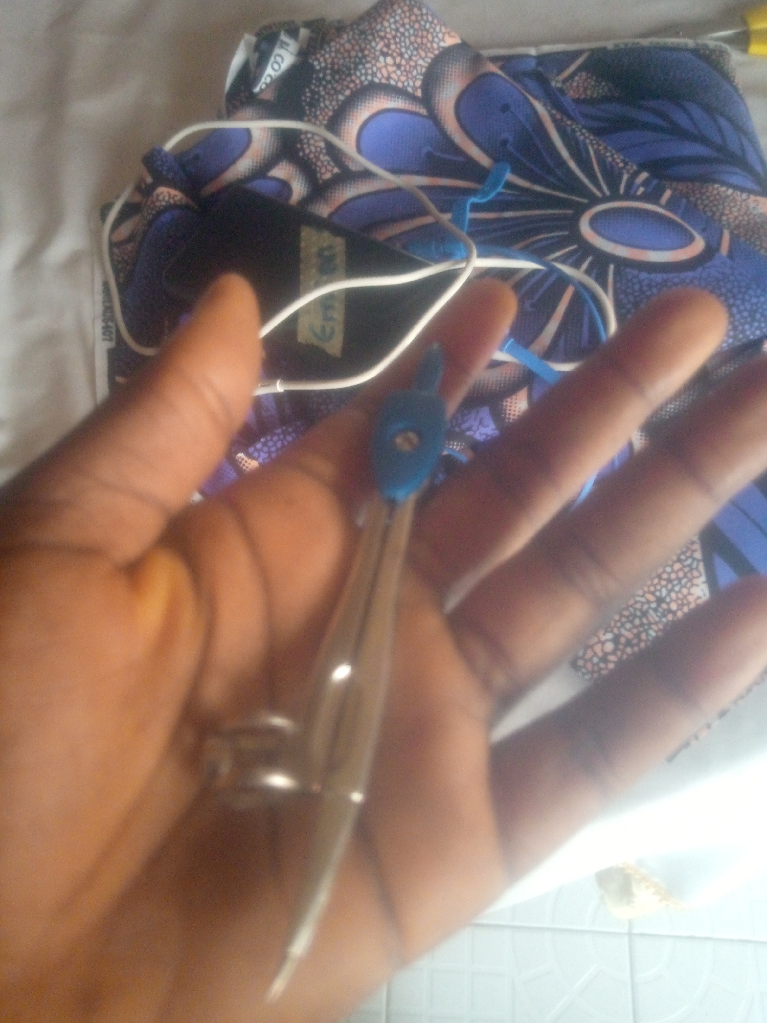 A close-up, dimly lit, overhead shot shows a person's hand holding a metal compass with a blue tip. The compass is positioned diagonally across the image, with its needle pointing towards the bottom left. The hand is a light brown color, with visible fingers and palm. In the background, a piece of fabric with a blue, purple, and pink floral pattern is visible, along with some white material. The scene appears to be indoors. There is no information to indicate this image is located in Jauro Yinu, Nigeria.