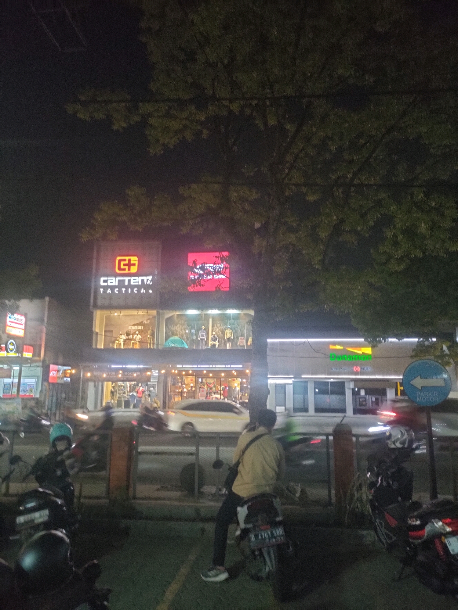 This is a night-time street-level, eye-level shot of a commercial building complex in Bandung City, Indonesia. The building features multiple businesses with illuminated signage. Prominently displayed is a sign for "Cartenz Tactical," featuring a red "G+" logo above the text. Next to it, a large, vibrant red sign displays what appears to be abstract graphics or text. Another business, "Danamon," is visible with its green and orange logo. The foreground shows a street with several motorcycles, some parked and some in motion, with their headlights and taillights creating light streaks. One person is seated on a motorcycle, wearing a light yellow jacket and a black bag over their shoulder. Another person is visible on a different motorcycle, wearing a blue helmet. The environment is characterized by artificial lighting from the businesses and streetlights, casting a low-light, nighttime ambiance. A blue sign with white text and an arrow pointing left, reading "PARKIR MOTOR," indicates a motorcycle parking area. Trees with dense foliage are visible in the upper portion of the frame, obscuring the night sky.