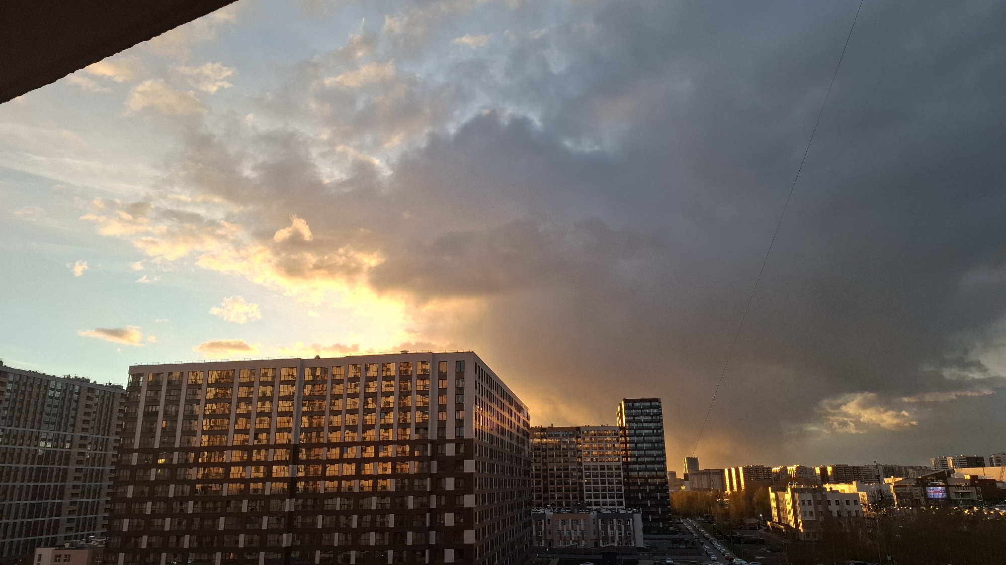 The image displays a dramatic urban landscape at either sunrise or sunset, characterized by a striking contrast in atmospheric conditions. The left side of the sky is bright with warm, golden-orange light reflecting off scattered clouds against a pale blue background, indicating direct sunlight from a low angle. This intense golden light bathes the windows of a prominent, multi-story modern building in the central and left foreground, making its facade glow.
In stark contrast, the right half of the sky is dominated by heavy, dark gray storm clouds, suggesting a weather front or a localized storm. A thin, dark power line or cable stretches diagonally across this darker expanse of sky. Below the horizon, numerous contemporary buildings populate the urban setting, characteristic of a large city like Saint Petersburg, Russia. Some buildings in the midground and background also catch the golden light on their upper sections, while others are silhouetted against the darker sky. A street with faint outlines of cars and some leafless trees is visible at the bottom of the frame, hinting at an active city environment.
No people are visible, and there is no discernible text in the image. The scene captures a powerful natural phenomenon over a man-made environment, emphasizing the interplay of light, shadow, and impending or receding weather.