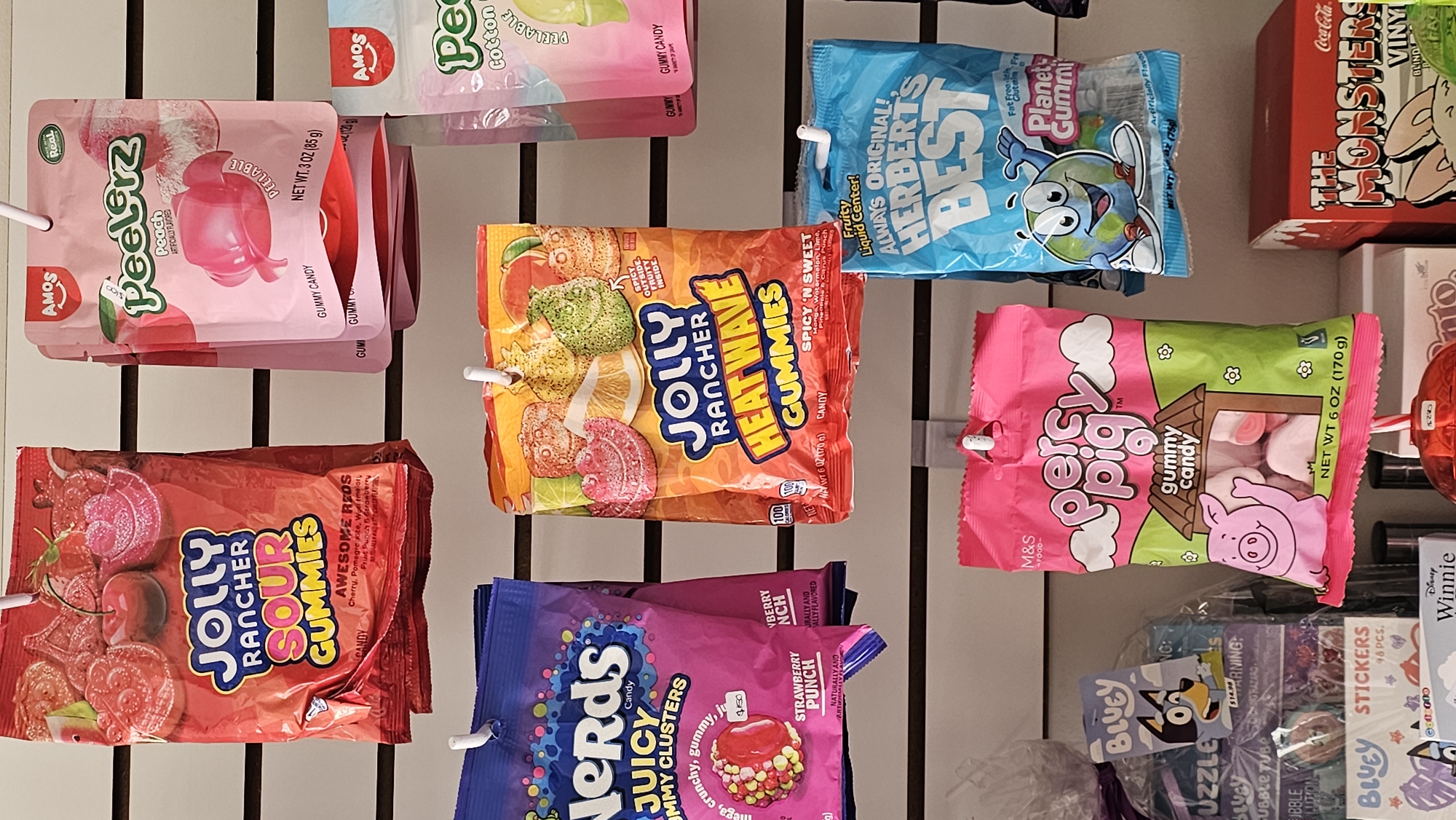 This is an eye-level shot of a retail display showcasing various bags of candy and novelty items, likely in a store or supermarket aisle. The products are hung neatly on white hooks against a white slatwall or pegboard. The lighting is bright and even, indicating an indoor setting.
On the top row, from left to right, are pink bags of "AMOS Peelerz Peach" gummy candy, described as "PEELABLE" and "ARTIFICIALLY FLAVORED," weighing "NET WT. 3 OZ (85 g)." Next to them, partially visible, is an "AMOS Peach Cotton Candy" bag. To the right, a blue bag features "Planet Gummi" and "ALWAYS ORIGINAL! HERBERT'S BEST" gummy candy with a "Fruity Liquid Center!" and marked as "Fat Free! Gluten Free!" Above these, a red "Coca-Cola THE MONSTERS VINYL BLIND BOX" is visible.
The middle row displays a red bag of "JOLLY RANCHER SOUR GUMMIES" with "AWESOME REDS" flavors like "Cherry, Pomegranate, Watermelon, Fruit Punch & Cranberry." Adjacent to it is an orange bag of "JOLLY RANCHER HEAT WAVE GUMMIES," highlighting "SPICY OUTSIDE FRUITY INSIDE" and "SPICY 'N SWEET" flavors including Orange, Watermelon, Lime, Pineapple & Citrus Punch, weighing "NET WT 6 OZ (170g)." To the right, a pink and green bag of "Percy Pig gummy candy" from "M&S FOOD-" is shown, also "NET WT 6 OZ (170g)."
The bottom row features a purple bag of "Nerds JUICY GUMMY CLUSTERS" in "STRAWBERRY PUNCH" flavor, described as "Mega, crunchy, gummy." Below the Percy Pig candy, various "BLUEY" branded items are visible, including "STICKERS 48 PCS." and a "BUBBLE TUBE" with a partial "WARNING: CHOKING HAZARD" label, priced at "$1.50". A "Disney Winnie" item is also partially visible on the far right.
No people or specific activities are depicted, focusing solely on the displayed products. The vibrant packaging and variety suggest a selection aimed at children and candy enthusiasts. The context provided specifies the location as San Bernardino Tlaxcalancingo, Mexico, though no direct visual cues in the image confirm this.