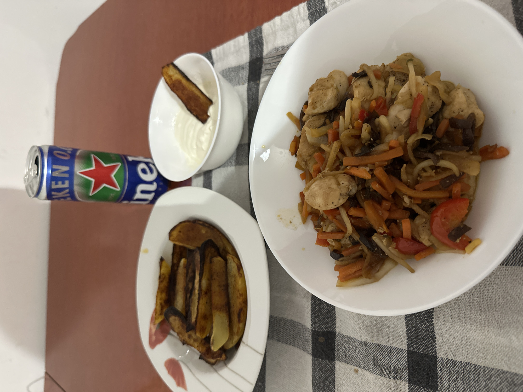An overhead shot shows a casual meal set on a table covered with a black and white checkered cloth.
The main subject is a large white oval bowl filled with a stir-fried dish. This dish contains pieces of cooked chicken, julienned carrots, sliced red bell peppers, and other vegetables that appear to be onions, mushrooms, or possibly thin noodles. The ingredients are mixed together and seasoned with visible specks of pepper.
To the left of the main bowl, a smaller white plate holds a serving of potato wedges or rustic-cut fries, which appear to be golden brown and slightly crisp. Above this plate, a small white bowl contains a creamy white sauce, possibly sour cream or mayonnaise, with a single elongated, browned vegetable piece (possibly fried eggplant or zucchini) resting in it.
Behind the smaller bowl and plate, a blue and silver Heineken 0.0 beer can is visible, standing upright. The can clearly displays the "HEINEKEN 0.0" branding and the iconic red star logo.
The background shows a plain, reddish-brown surface, likely a table or countertop. The overall setting suggests a relaxed, indoor dining experience, possibly at home. The time of day and specific location are not discernible.