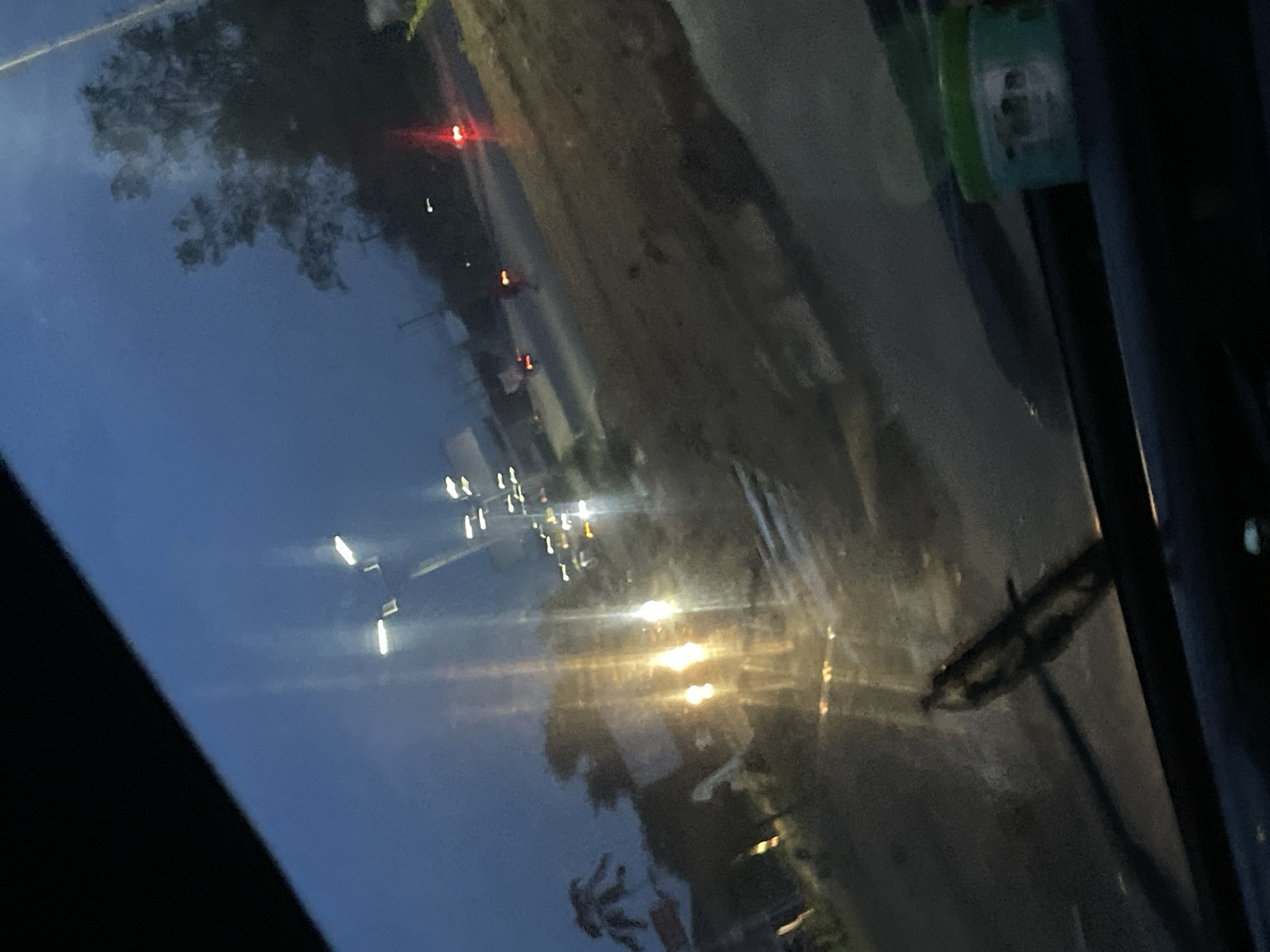 The image depicts a view through a windshield at dusk or dawn, likely in Jos, Nigeria. Streetlights are visible, some with a yellow-orange glow, illuminating a road with patchy, wet surfaces, suggesting recent rainfall or a damp environment. The scene is somewhat blurry, a characteristic often associated with low light conditions and movement. Trees with dark foliage are silhouetted against a pale blue and darkening sky. There are signs of traffic, with distant red taillights and white headlights visible on the road, indicating vehicles in motion. The windshield itself shows streaks and reflections, further contributing to the indistinct quality of the image. In the lower right corner, a green lidded container is visible on the dashboard, partially obstructing the view of the road ahead. A windshield wiper blade is also in frame, positioned across the lower left portion of the windshield. The overall impression is of a fleeting glimpse of a street scene in Jos during twilight hours, possibly from inside a moving vehicle.