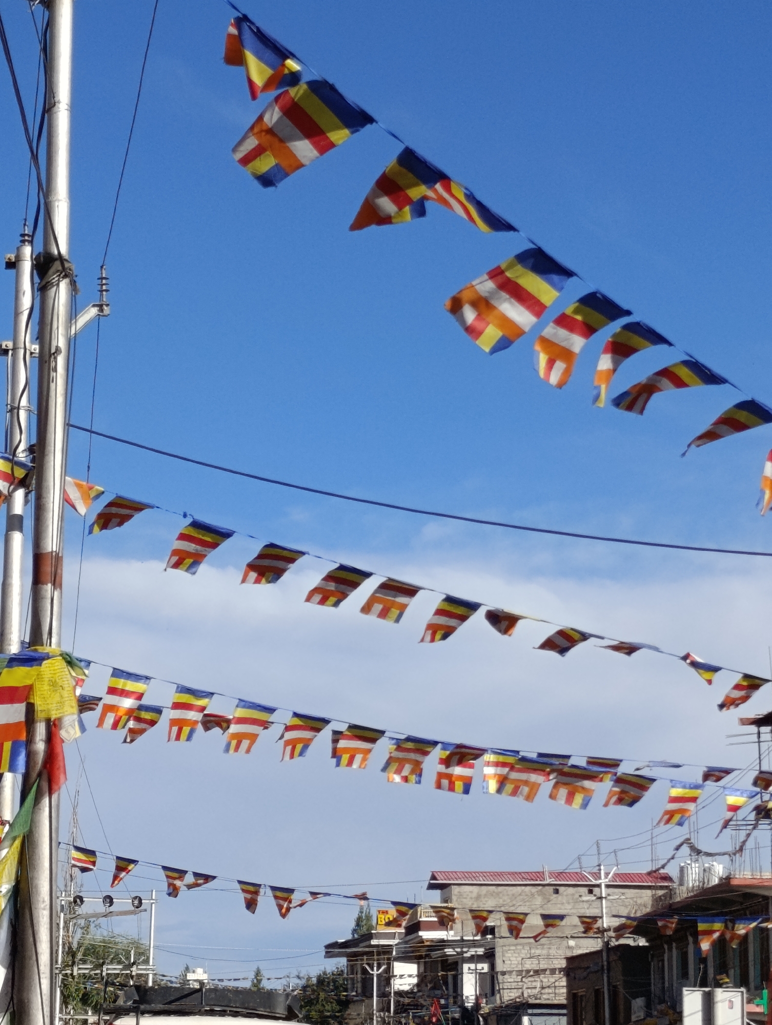The image displays strings of prayer flags strung across a clear blue sky, with hints of white clouds. The flags are colorful, featuring a repeating pattern of horizontal stripes in blue, yellow, red, white, and orange. They are hung from wires or ropes, with one main visible pole on the left side of the frame. In the background, buildings are visible, suggesting an urban or semi-urban environment. The style of the flags and the architecture of the buildings are consistent with locations in Ladakh, India, such as the region around Thiksey Monastery. There are no people clearly visible in the foreground or mid-ground of the image. The scene appears to be a bright, sunny day.
