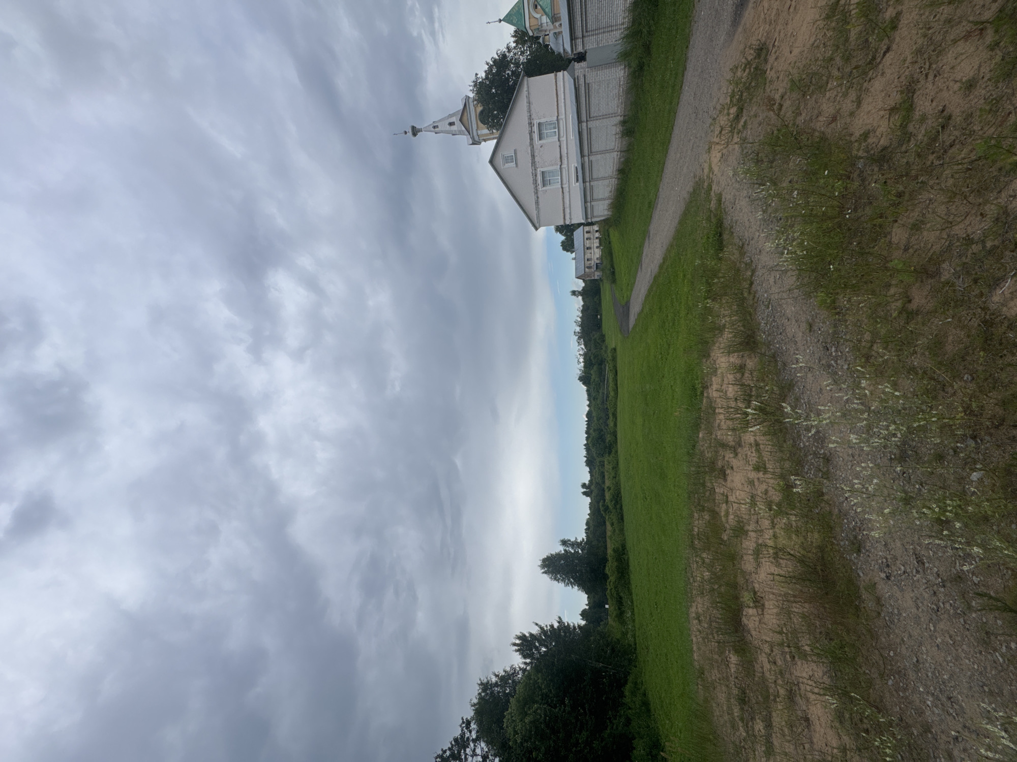 The image depicts a pastoral scene on an overcast day. A gravel path curves from the foreground towards a cluster of buildings on the right, which appear to be part of a religious complex. One building, painted white with a pale yellow steeple topped with a small golden dome, is prominent. A low white brick wall borders this building and extends to the right. Beyond the path and wall is a vast expanse of bright green grass. To the left of the path, the terrain transitions to a sandy area with sparse vegetation before meeting a dense line of dark green trees. Further in the distance, more trees form a natural horizon. The sky is filled with heavy, grey clouds, with a faint hint of blue sky peeking through in the far distance. The overall impression is one of quiet rural life in the settlement of Rassvet, Russia.