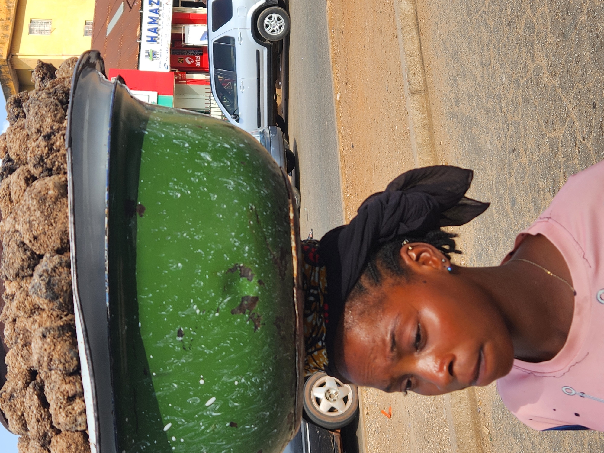 A woman carries a large green basin filled with what appears to be roasted plantains. The basin has a black rim. The woman wears a pink shirt and a black headscarf. She has multiple ear piercings. In the background, a white SUV is parked next to the road. Buildings and signs are visible in the distance, suggesting an urban setting. The location is in the city of Unknown, Unknown.