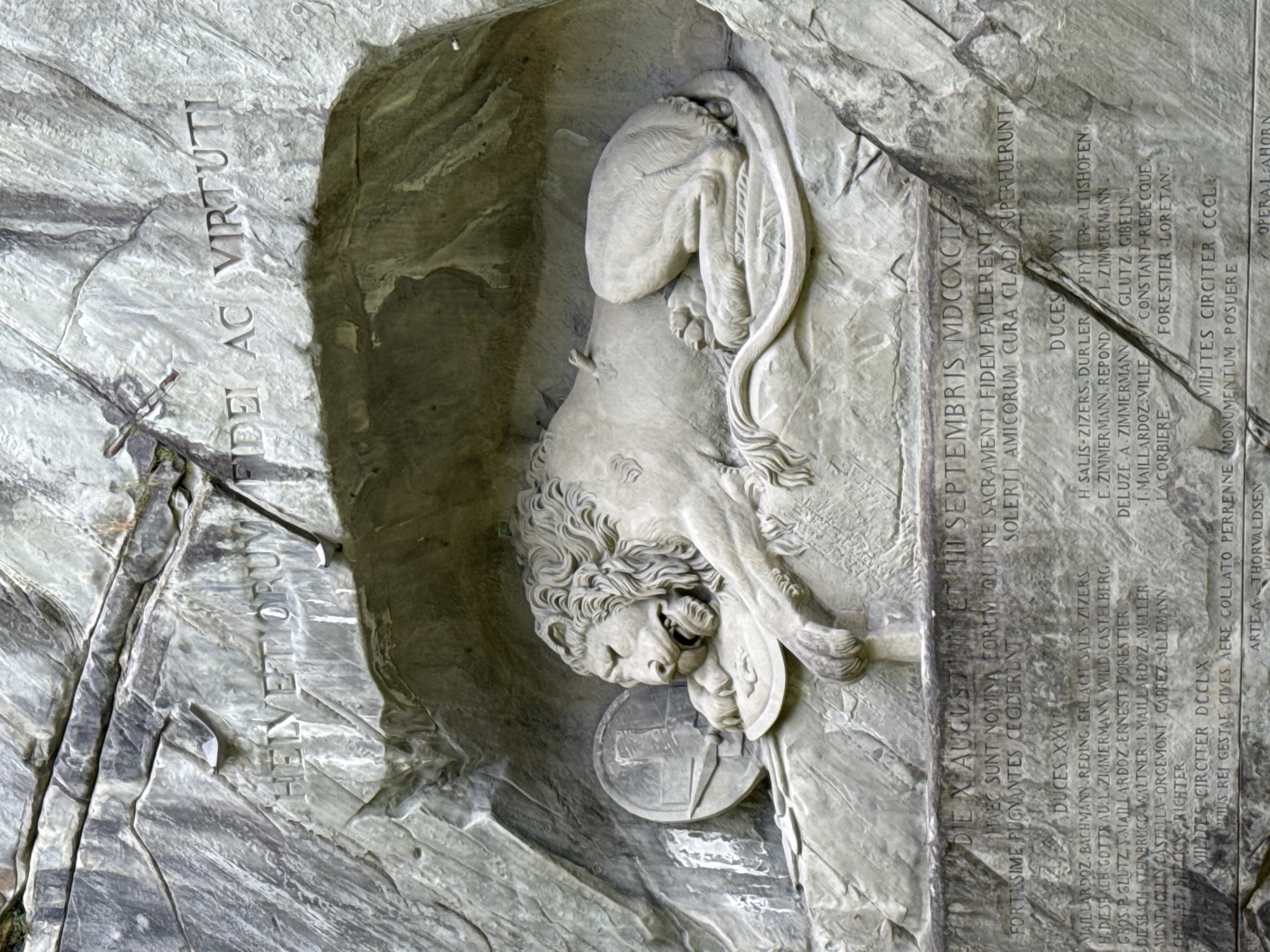 The image shows the Lion Monument in Lucerne, Switzerland. It is a rock relief sculpture carved into a cliff face. The central subject is a dying lion, mortally wounded by a spear, clutching a shield with the fleur-de-lis of the French monarchy. The lion is depicted in a state of agony, with its head lowered and mouth open. To the left of the lion is a shield with a cross, representing Switzerland. Above the lion, carved into the rock, is the Latin inscription "HELVE TIORUM FID EI AC VIRTUTI" which translates to "To the loyalty and bravery of the Helvetii". Below the lion, a long list of names is carved into the rock, commemorating Swiss guards who died during the French Revolution. The monument is situated in a natural rock crevice, with rough-hewn stone surrounding the sculpted figures and inscriptions. The overall tone of the monument is somber and commemorative.