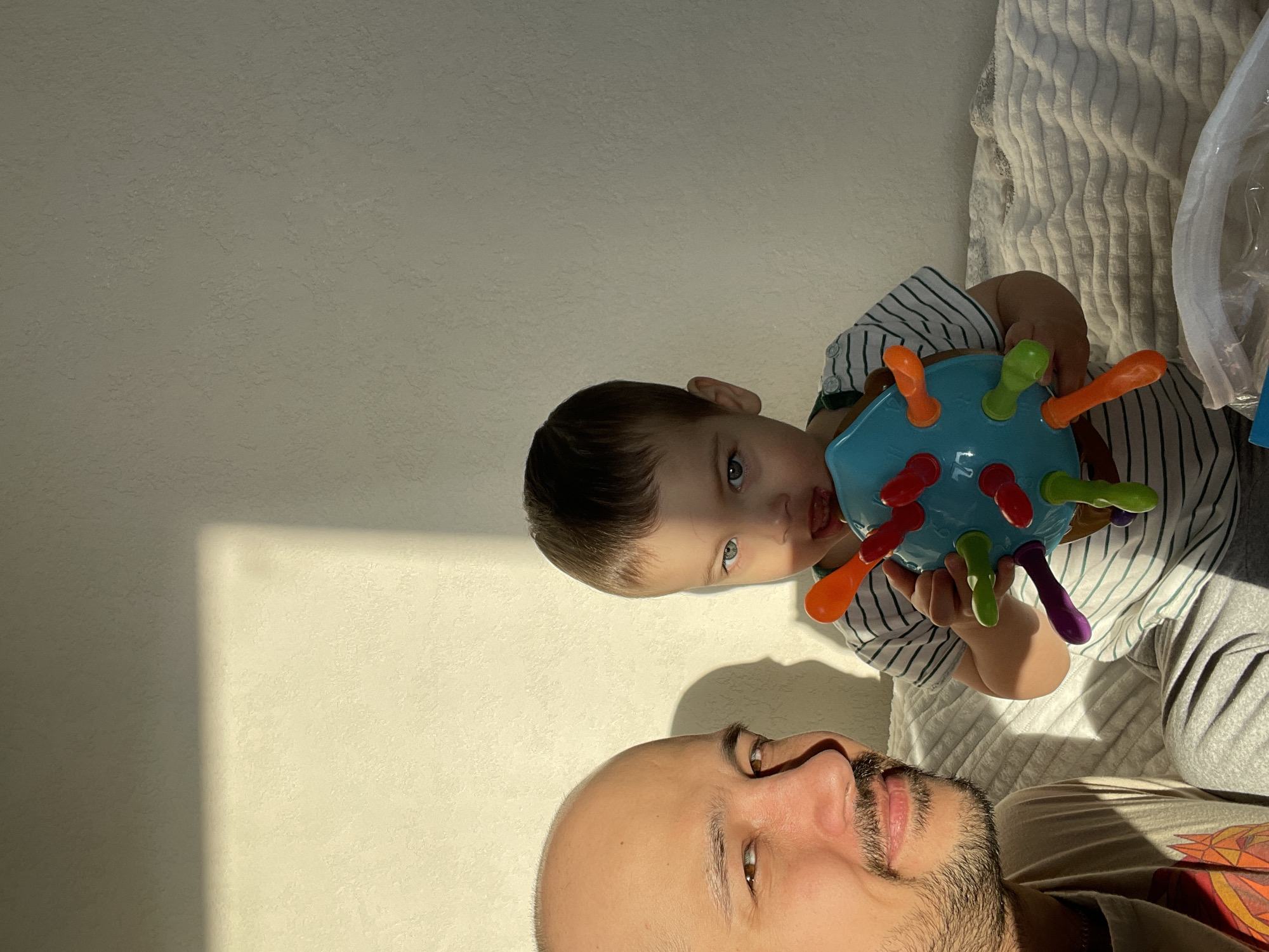 The scene is situated indoors, purportedly in a residential location within Izhevsk, Russia. It features an adult male and a young child. The adult male, visible in the left foreground, has a shaved head, dark stubble, and is smiling while looking towards the camera. He wears a light-colored t-shirt with a graphic design partially visible. The child, positioned to the right of the man, has dark hair and light-colored eyes. The child is holding a light blue, spiky sensory toy with red, orange, green, and purple flexible pegs, one of which is partially in the child's mouth. The child wears a white and dark-striped t-shirt. Both individuals are seated on a surface covered by a white, textured blanket, which extends across the lower portion of the frame. A partially visible white plastic bag is located in the bottom right. The background is a plain, light-colored textured wall. Strong, directional natural light from the left illuminates the subjects, casting sharp shadows on the wall behind them.