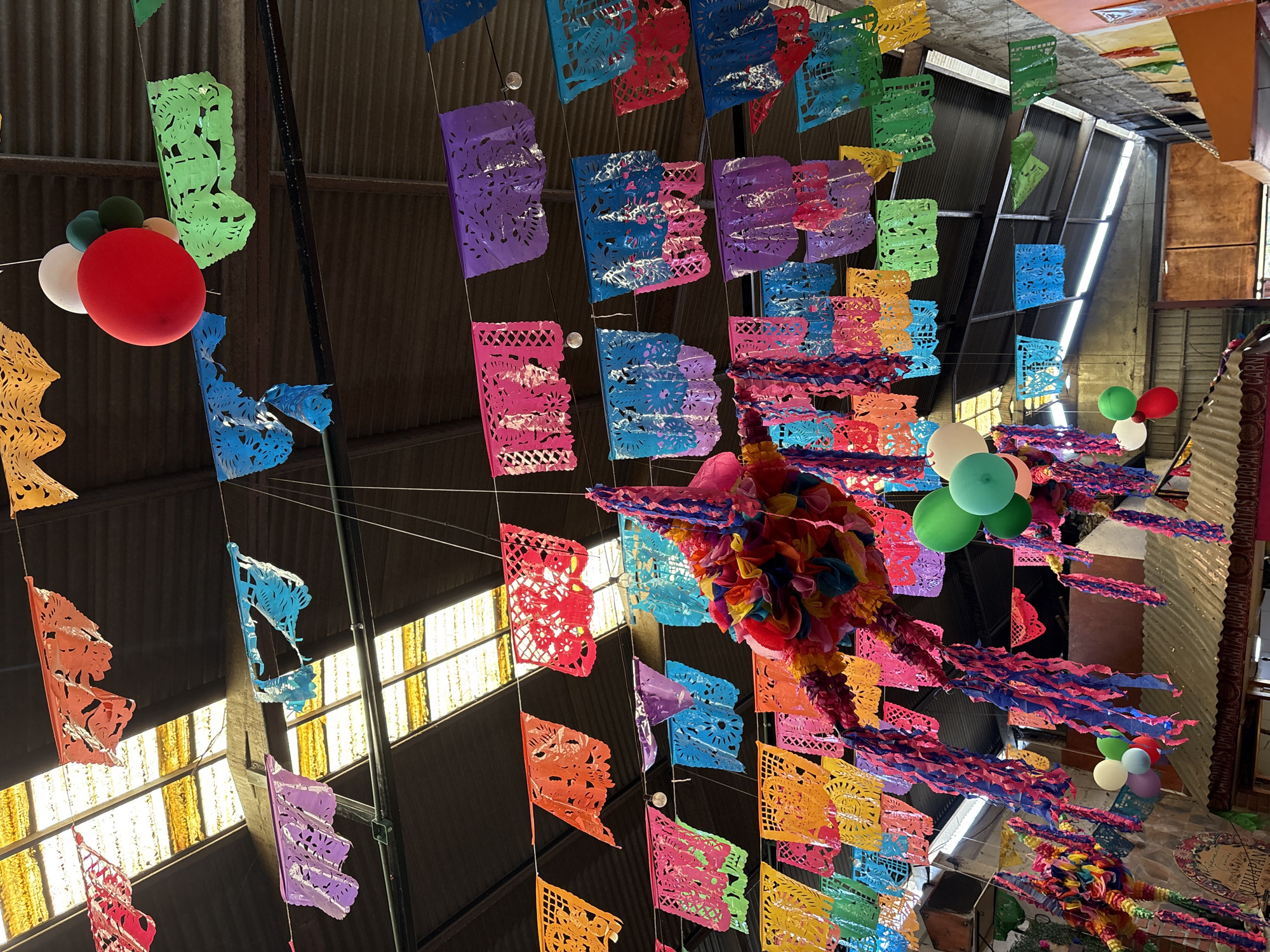The image captures an indoor space adorned with colorful Mexican decorations. Cut paper banners in various colors are strung overhead. Balloons are clustered in several locations. A piñata hangs centrally. The location is Ocotepec, Mexico, as inferred from the setting and decorations.