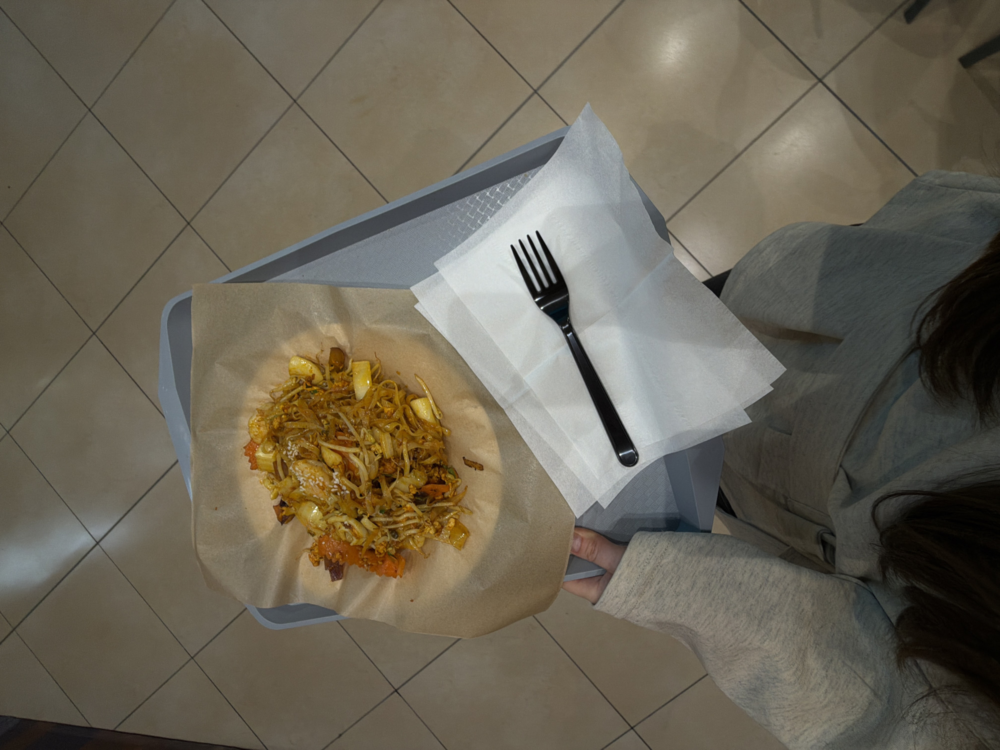 An overhead shot depicts a meal on a gray tray held by a person in a light gray sweatshirt. The meal, a stir-fried dish with bean sprouts and possibly meat, is served on brown paper. Beside the dish is a stack of white napkins with a black plastic fork on top. The person’s dark hair is visible in the lower right of the frame. The background shows a tiled floor.
The city of Khimki, Russia, is not identifiable in the image.