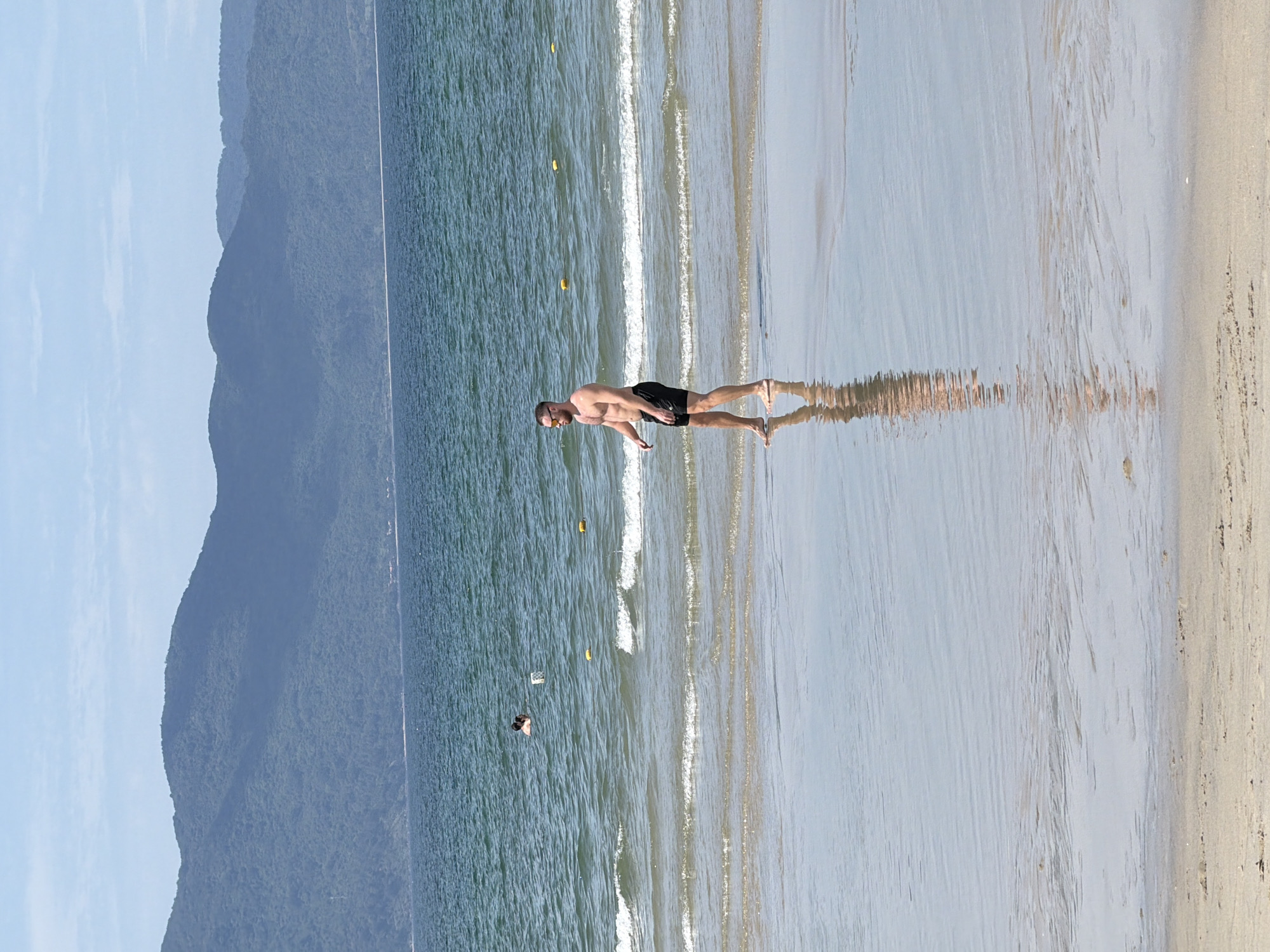 The image depicts a beach scene in Đà Nẵng, Vietnam. In the foreground, a man with light skin and short brown hair is walking along the wet sand at the edge of the water. He is wearing black swim trunks and sunglasses. His reflection is visible on the wet sand. In the middle ground, the ocean is calm with gentle waves rolling onto the shore. Two individuals are visible in the water in the distance. Yellow buoys are scattered across the water. In the background, a range of lush green mountains forms a natural backdrop against a pale blue sky with scattered clouds. The overall scene conveys a serene and sunny day at the beach.