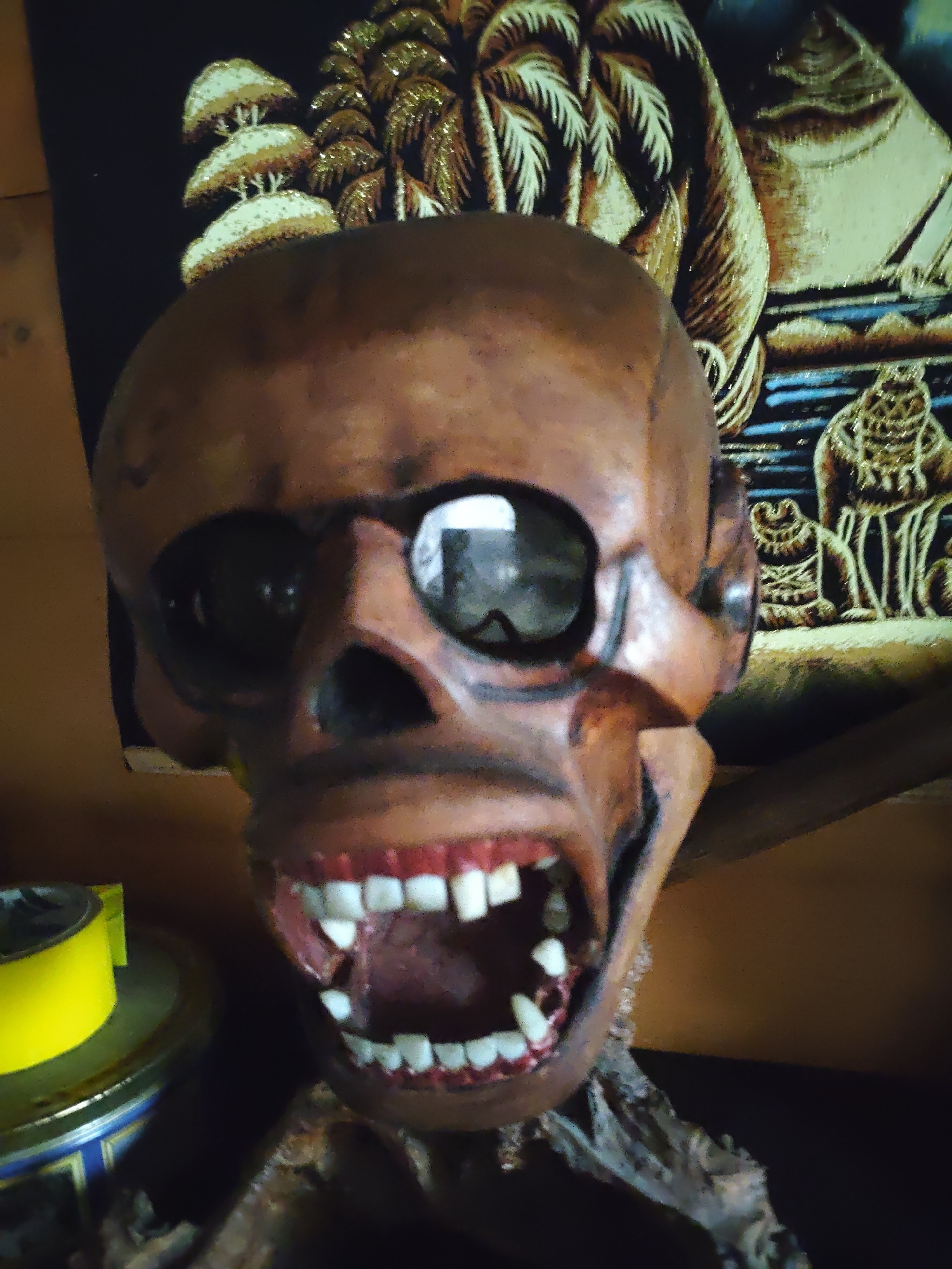 The image depicts a close-up of a decorative skull with sunglasses and an open mouth revealing teeth. The skull is brown and appears to be made of a composite material. The background includes a tapestry-like object with imagery of trees and figures. A yellow tape dispenser rests on a surface to the left of the skull. The scene is located in Боровково, Russia.