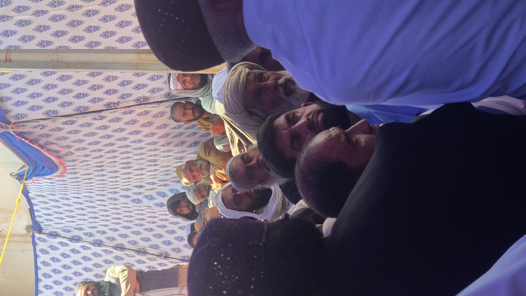 The image shows a gathering of men under a tent with a white and blue patterned ceiling. Most of the men have beards and are wearing traditional Muslim attire, including hats and turbans. Some men are wearing Western-style suits. The faces are mostly middle-aged to elderly. The location may be Panjpai, Pakistan, though this cannot be verified with the image alone. The purpose of the gathering is unclear from the photo, but the context suggests a community event or a religious gathering.