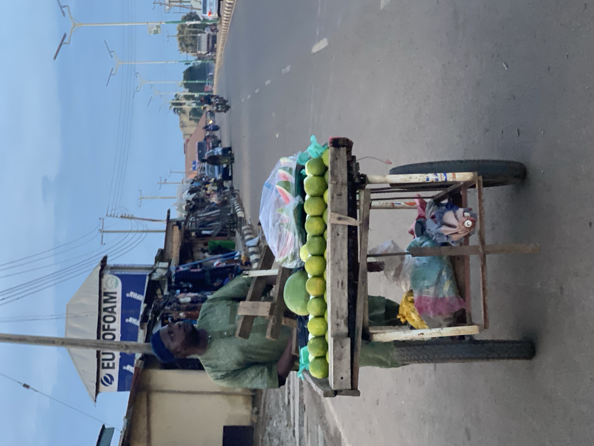 This image captures a street scene in Kafanchan B, Nigeria, under an overcast sky. In the foreground, a man of dark complexion, wearing a green traditional garment and a dark cap, stands behind a wooden pushcart. The cart, equipped with two dark wheels, displays produce on two levels. The top shelf features a neat row of approximately twelve bright green limes or oranges, a large green watermelon, and several clear plastic bags containing sliced watermelon. A light blue plastic bag is also on the top surface. The lower shelf holds multiple colorful plastic bags, including yellow, pink, and blue, containing various items.
Behind the man and his cart, to the left, a series of informal market stalls and structures line the roadside. Above these structures, a blue signboard partially obscured by a utility pole displays the white text "EU OFOAM," indicating a business. The utility pole is laden with numerous overhead wires. Several individuals are visible amidst these stalls.
Extending into the midground and background, an asphalt road with white dashed lane markings leads away from the viewer. A concrete median barrier separates the two directions of traffic. Numerous vehicles, including cars, motorcycles, and a white truck, are observed traveling on the road. Tall streetlights with solar panel tops line the median. The background features more green foliage and additional utility poles and power lines against the light blue-grey sky.