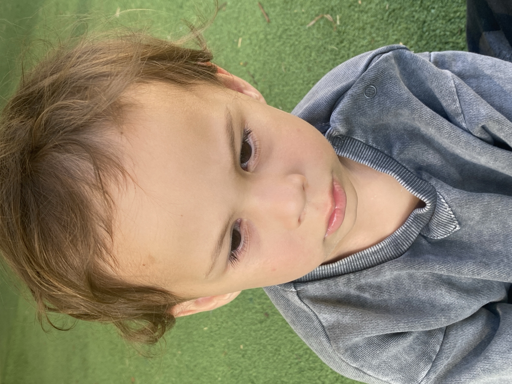 A close-up, eye-level shot shows a young child with light brown hair and fair skin looking downwards with a slightly pouty expression. The child is wearing a grey, long-sleeved t-shirt with a ribbed collar and snaps on the shoulder. The background is a blurred green surface, possibly artificial turf, consistent with an outdoor recreational area or playground. There is no indication of the location being Ashdod, Israel.