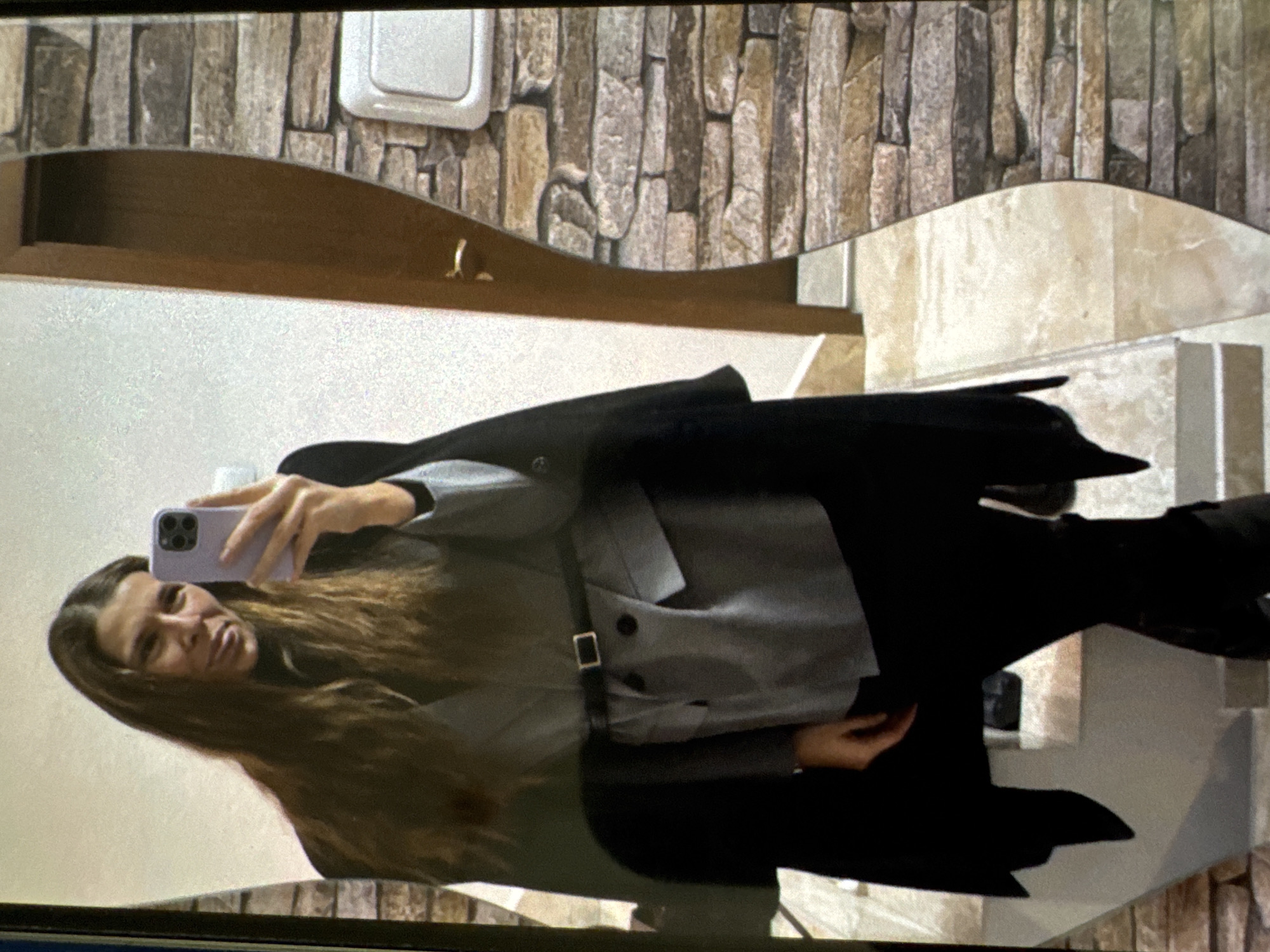 The image depicts a woman taking a mirror selfie within an indoor setting.
**Person:**
A female subject with long, straight brown hair is positioned centrally in the frame. She has a faint smile and is looking towards the camera of a light purple smartphone, which she holds with her right hand.
She is dressed in a black turtleneck top, layered with a grey blazer. The blazer features a single button closure, lapels, and two flap pockets on the front. A thin black belt with a rectangular silver-toned buckle cinches the blazer at her waist. Over the blazer, she wears a black, open-front cape or coat with wide, flowing sleeves/panels that extend down her sides. She is also wearing black pants or leggings and black boots, the lower portion of which are visible.
**Environment (reflected in the mirror):**
The mirror has an irregular, wavy-curved right edge (viewer's perspective) and a straight left edge. The reflected background reveals an interior space:
- To the left of the reflection (viewer's right in the image), there is a wall surfaced with decorative, stacked stone-like tiles in varied earthy tones of brown, grey, and beige. A white rectangular electrical outlet or light switch is mounted on this wall segment.
- To the right of the reflection (viewer's left in the image), a plain white wall is visible. A light brown wooden door with a gold-colored handle is partially open on this wall.
- Below the door and extending across the lower portion of the reflection, a staircase or multi-level floor is visible, featuring light-colored, possibly marble or tiled, steps and risers in shades of cream and beige.
**Location Context:**
The image provides no visual indicators or specific features to identify the location as the city of Vratsa, Bulgaria.