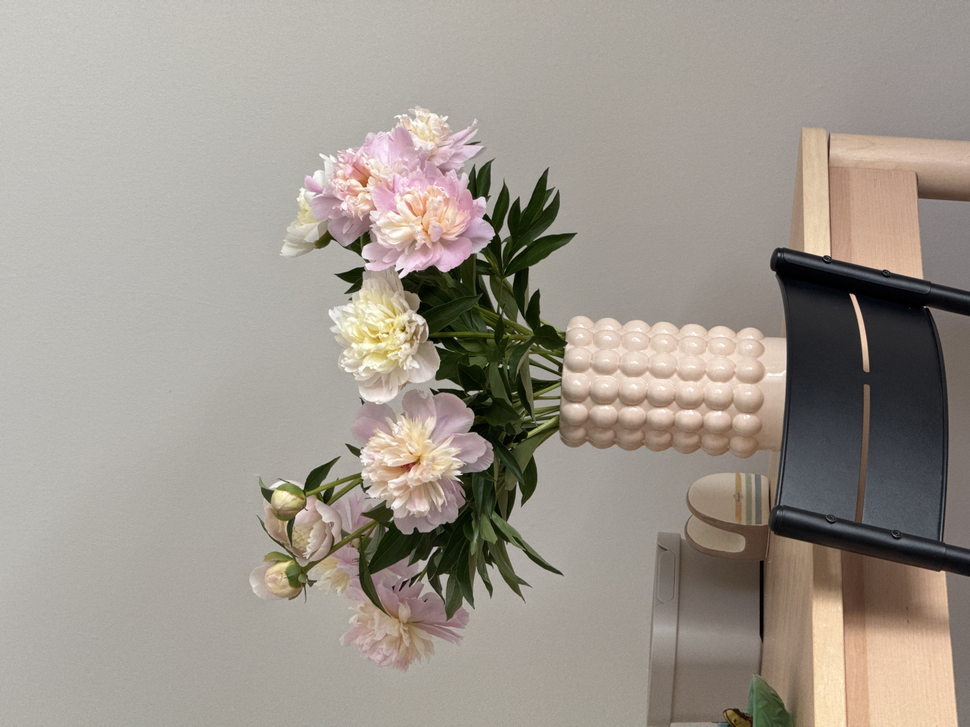 A close-up, eye-level shot shows a bouquet of pale pink and cream peonies in a textured, bubble-shaped light pink vase. The vase is placed on a light-colored wooden table. To the left of the vase, a light beige rectangular storage container with a lid and a small green bowl are visible. Further left, a brown and white striped object, possibly a napkin holder, sits on the table. A black chair with horizontal slats is positioned in front of the table, with its back to the viewer. The background is a plain, light gray wall. The scene is indoors, with natural light illuminating the table and flowers. There are no people present in the image. The specific location in Izhevsk, Russia cannot be determined from the image content.