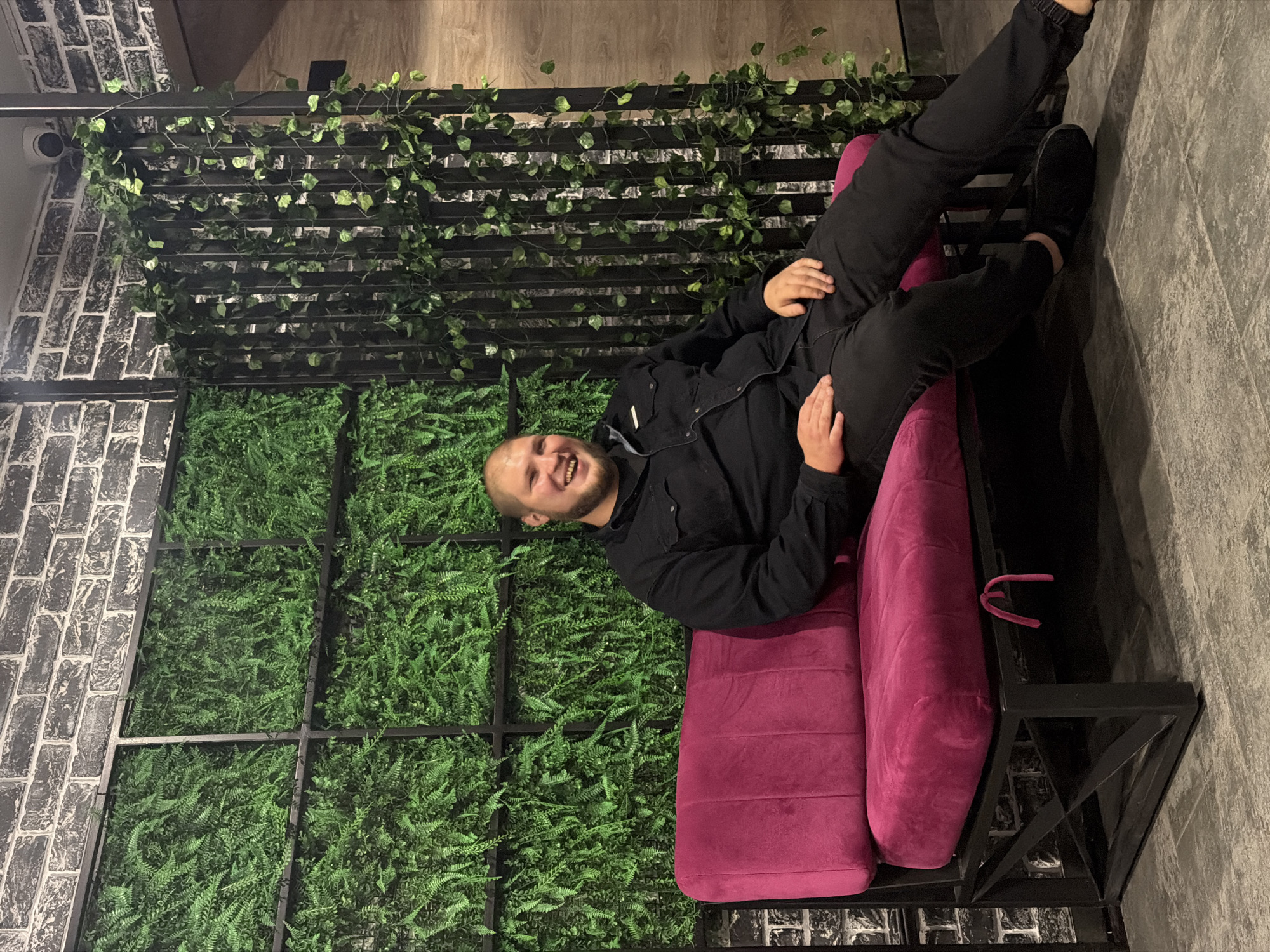 A Caucasian man with a bald head and a beard is sitting on a magenta sofa. He is wearing a black button-up shirt and black pants. The sofa is in front of a wall with a black brick pattern. To the left of the brick wall is a section of artificial ferns arranged in a grid. To the right of the brick wall is a black metal trellis covered with artificial ivy. The floor is tiled with a gray pattern. The man is smiling and looking at the camera. This scene is located in the city of Chaikovsky, Russia, likely in a cafe or a restaurant.