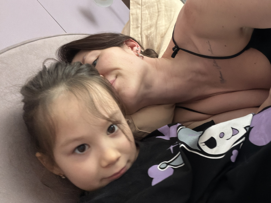 A young girl with brown hair and eyes looks directly at the camera, wearing a black shirt with cartoon figures in white and lavender. Behind her, a woman with dark hair and fair skin leans close, her face partially visible. The woman is wearing a black top with thin straps. A tattoo is visible on her upper chest/neck area. The background includes a pale lilac surface and a beige, rumpled fabric.
