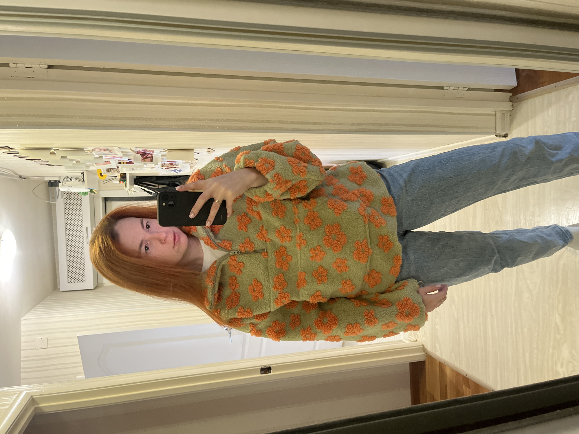 A young woman with long, reddish-brown hair stands in a hallway, taking a mirror selfie. She is wearing a light green, fuzzy quarter-zip pullover adorned with an all-over orange floral pattern. She is also wearing light blue denim jeans. The hallway features light-colored, vertical striped wallpaper and a wooden floor. To the left of the woman is a door, and to her right, the hallway continues with a doorway visible at the end. On the wall to the right of the woman, there are several framed pictures and other decorative items pinned to the wallpaper. The image appears to be taken indoors, possibly in a residential setting in Chaikovsky, Russia, as indicated by the provided metadata.