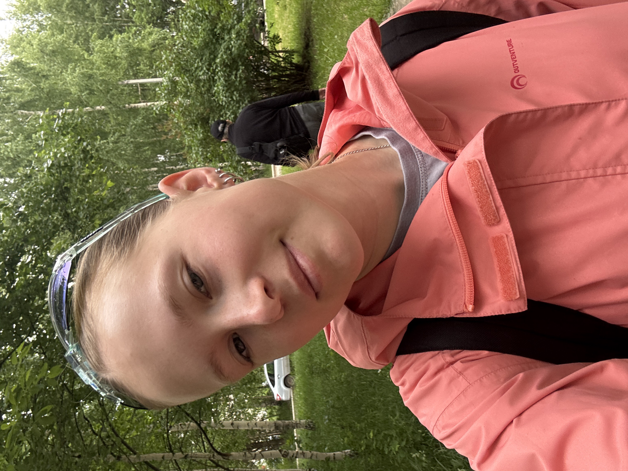 A young person, likely female, is featured prominently in a selfie, taken outdoors in what appears to be a park or green recreational area in Chaikovsky, Russia. She has light-colored hair, worn back, and is wearing sunglasses pushed up onto her head. Multiple small hoop earrings adorn her left ear, and a thin silver chain is visible around her neck. Her expression is soft and appears to be a slight, relaxed smile. She is wearing a salmon-pink or coral-colored outdoor jacket with a visible black backpack strap on her right shoulder. The jacket features the brand name "OUTVENTURE" with a circular logo on the upper right chest area.
In the blurred background, another person is seen from behind, wearing a black long-sleeved top and a black baseball cap. They appear to be walking or standing further down a path, surrounded by abundant green trees and grass. A white car is partially visible in the mid-ground. The lighting is bright and diffused, suggesting it is daytime, possibly an overcast or partly cloudy day, as there are no harsh shadows. The overall scene conveys a casual, outdoor activity.